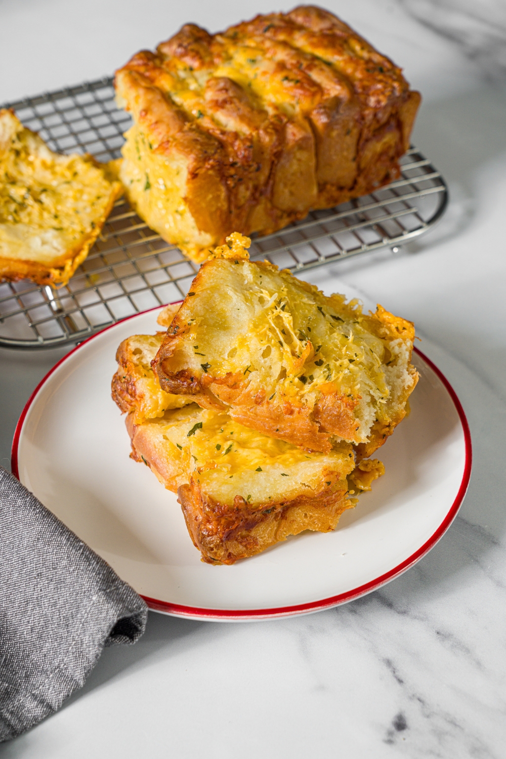 A white plate with two slices of pull apart cheesy garlic bread. The plate is on a marble counter with a drying rack of bread.