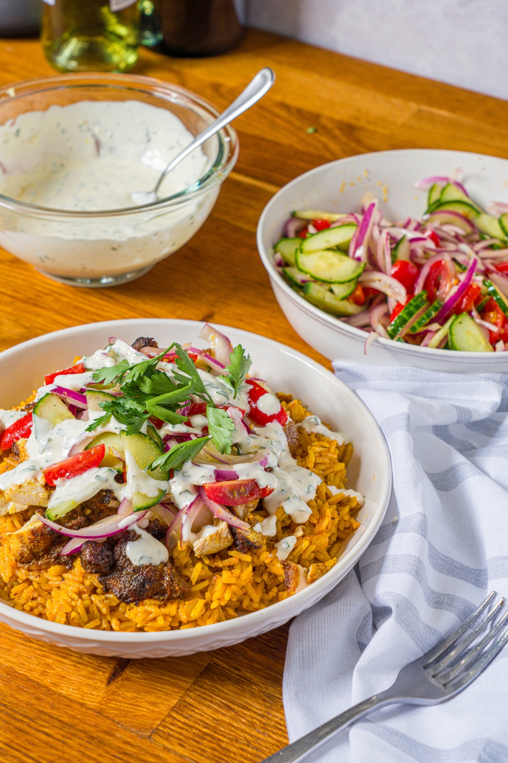 A white bowl with chicken shawarma bowl served with seasoned rice, chicken shawarma, onion salad, and garlic sauce. The bowl is on a wooden counter with a bowl of onion salad and bowl of garlic sauce.