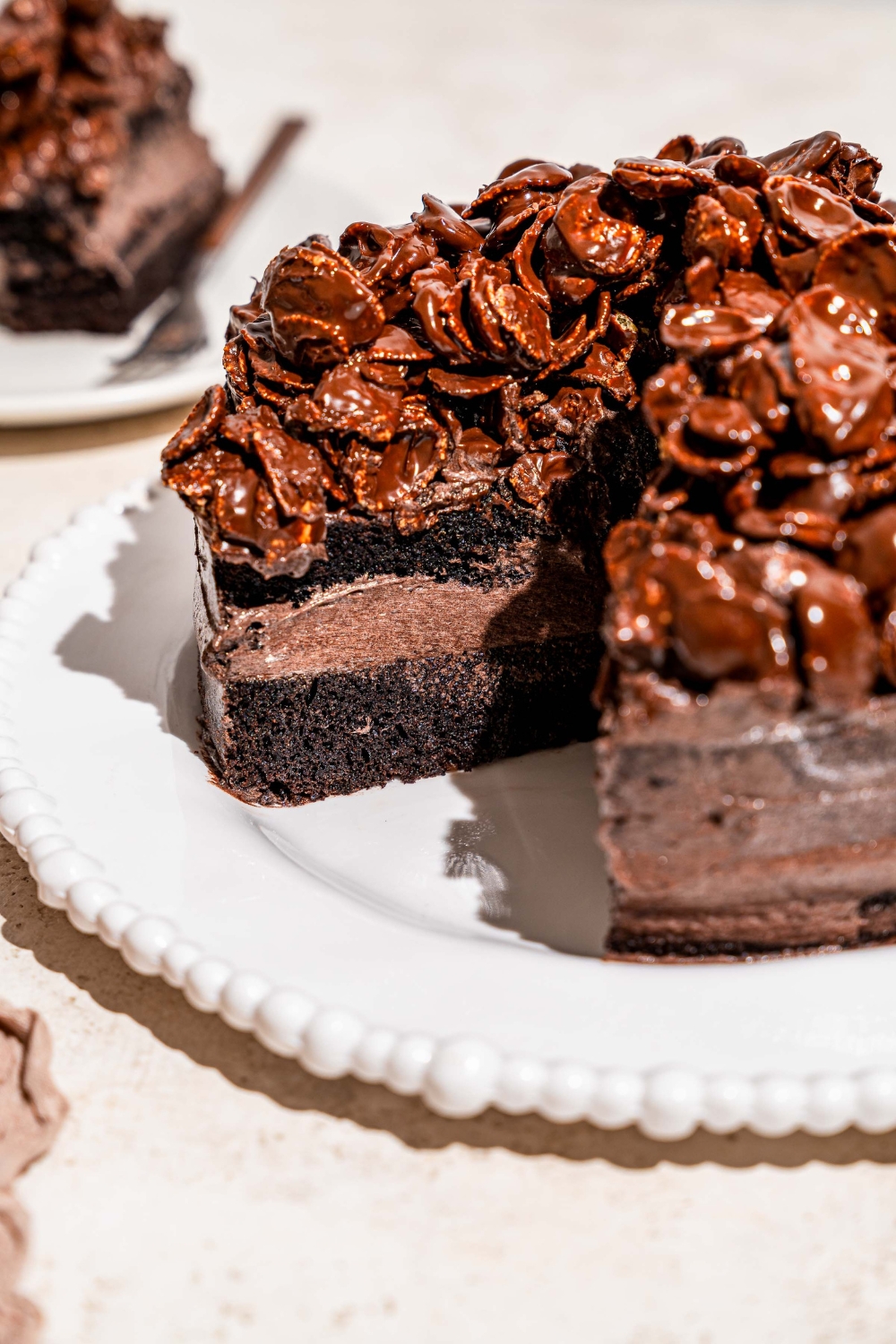 A white platter with a whole chocolate crunch cake topped with chocolate cornflake crunch. A slice is removed from the cake. The platter is on a tan counter with a plate with a slice of cake.