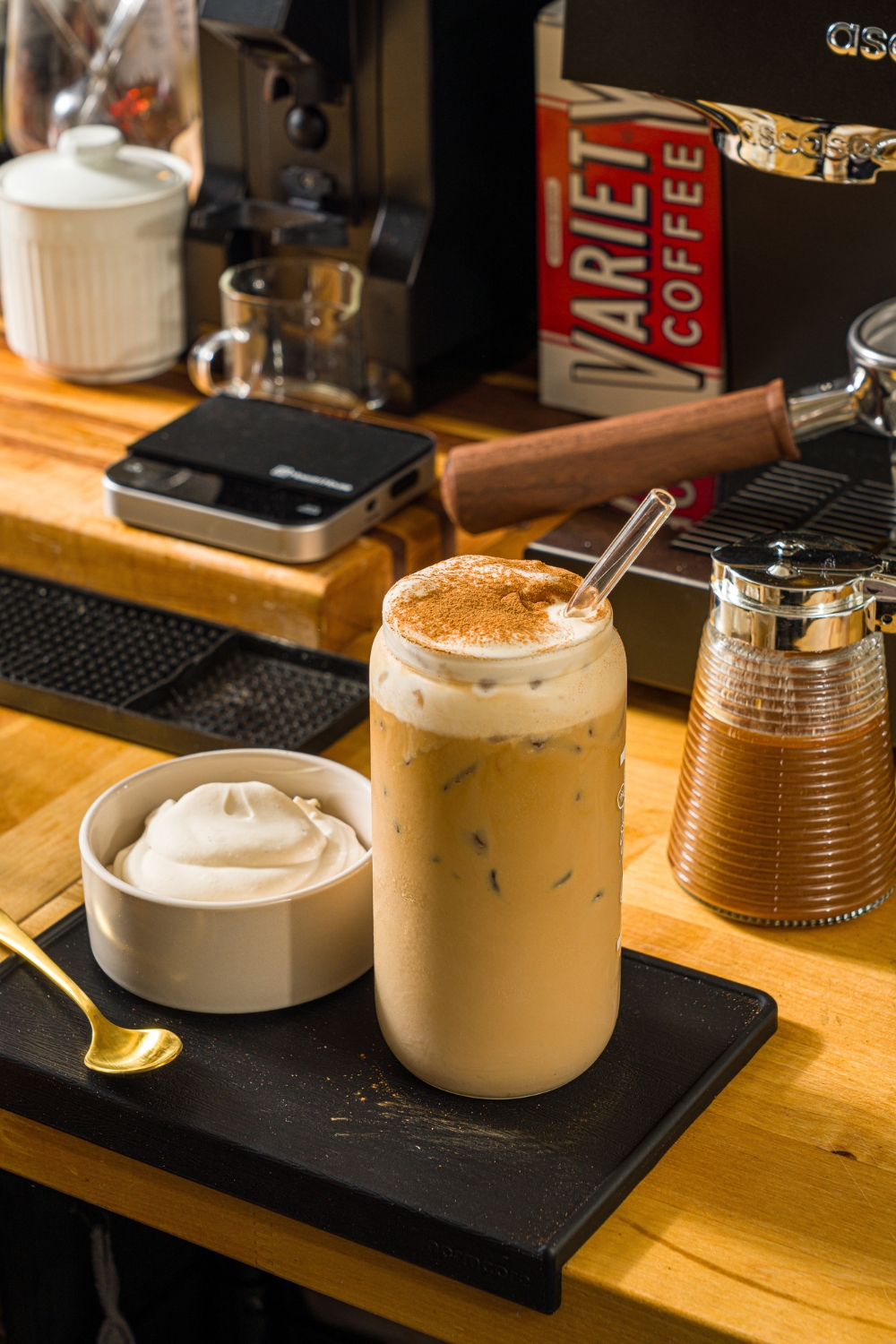 A tall glass with a glass straw with iced cookie butter latte topped with cold foam and cinnamon. The glass is on a wooden counter with a bowl of cold foam and an espresso machine.