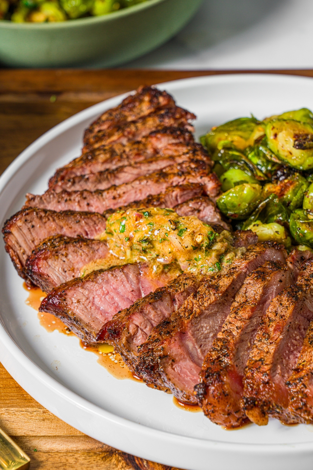 A white plate with sliced grilled steak topped with cowboy butter and a side of roasted brussels sprouts. The plate is on a wooden board.