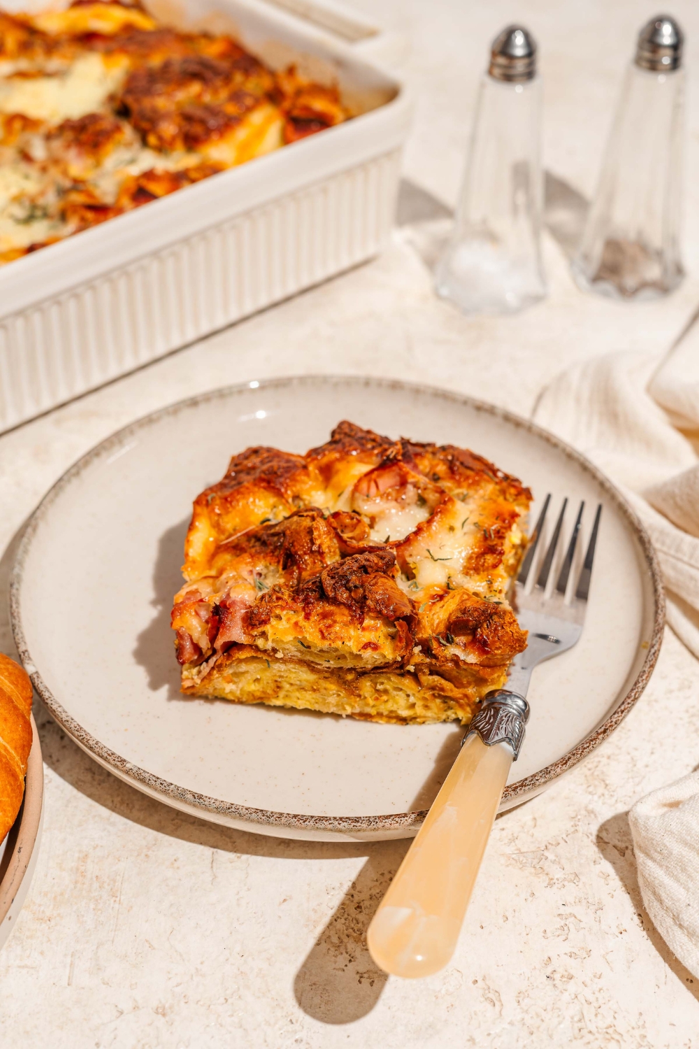 A white plate with a slice of croissant breakfast casserole with ham and cheese. There is a fork on the plate. The plate is on a tan counter with a baking dish of casserole and a salt and pepper shaker.