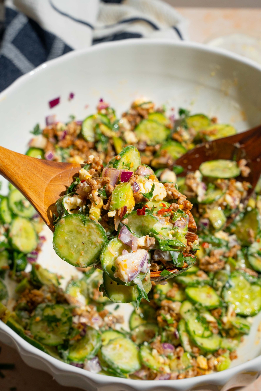 A close up of a salad tong with a scoop of high protein cucumber salad. There is a large bowl of. salad blurred in the background including cucumbers, feta cheese, seasoned ground beef, red onion, and avocado tossed in a greek yogurt dressing.