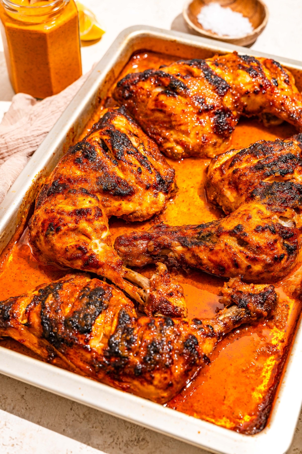 A sheet pan with roasted chicken cooked in peri peri sauce. The pan is on a tan counter with a jar of sauce.