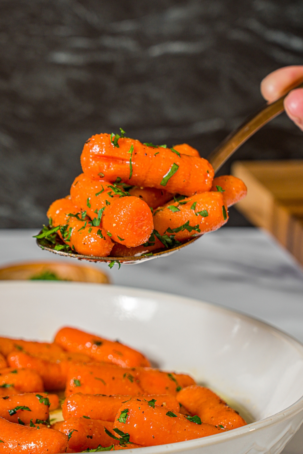 A close up of a spoon with honey roasted baby carrots garnished with fresh parsley. There is a bowl of carrots on a marble counter blurred in the background.