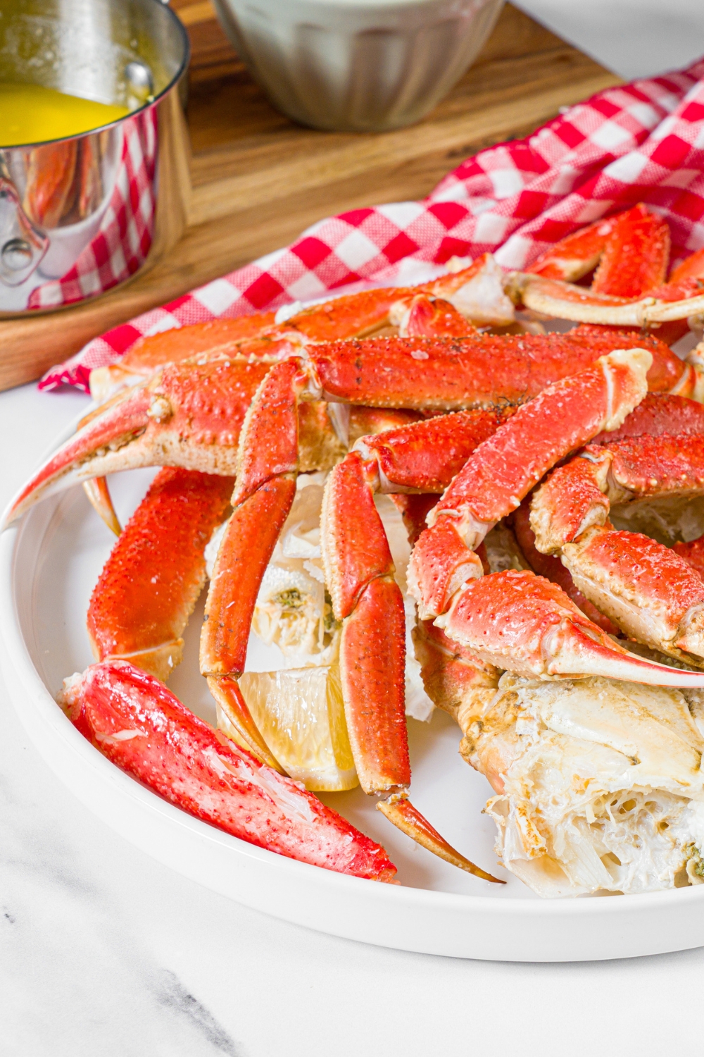 A white plate with steamed snow crab legs served with lemon. The plate is on a marble counter with a board of lemon butter sauce and a red checkered napkin.