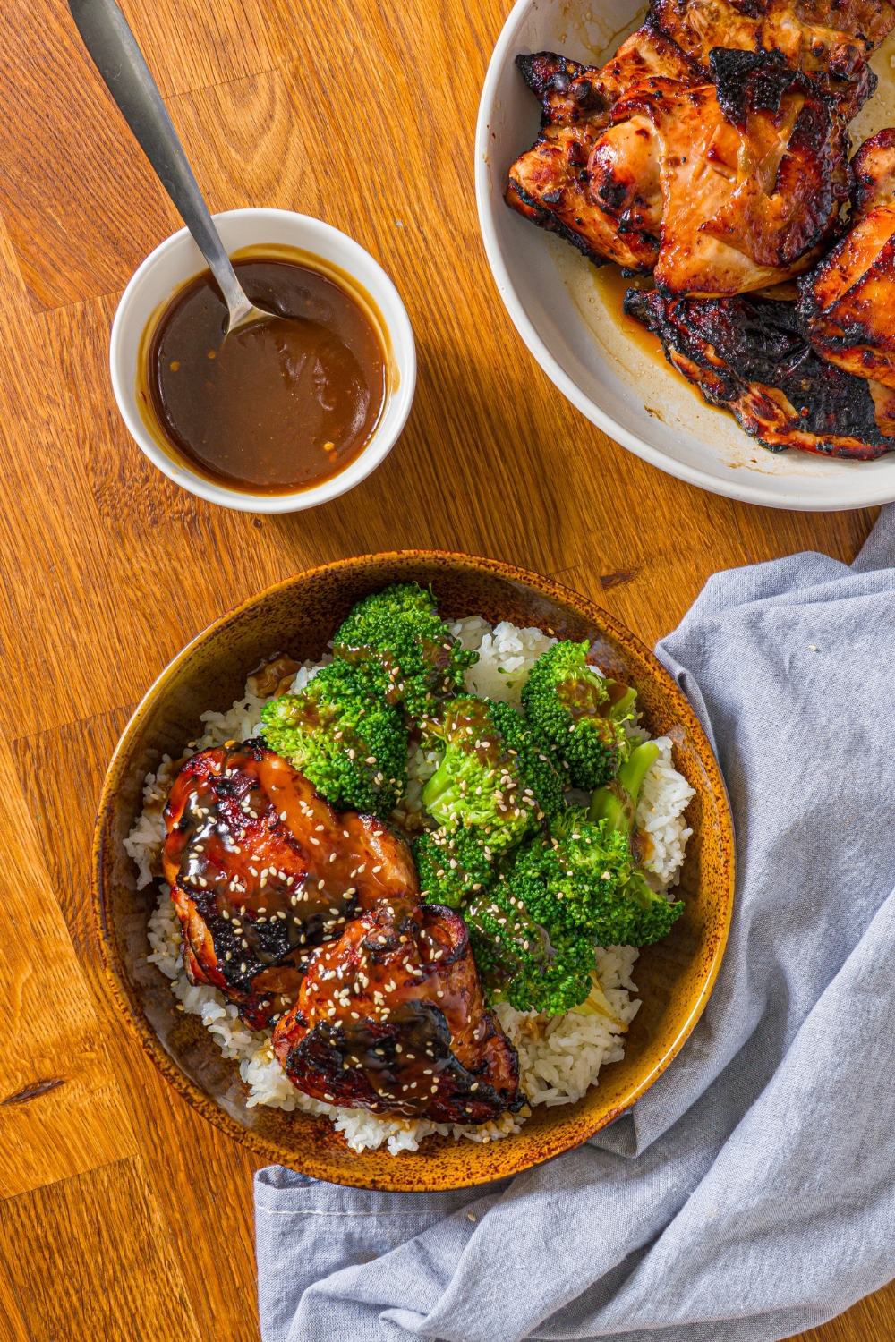 A bowl with chicken teriyaki served with broccoli and rice and garnished with sesame seeds. The bowl is on a wooden counter with a bowl of cooked chicken and small bowl of teriyaki sauce.