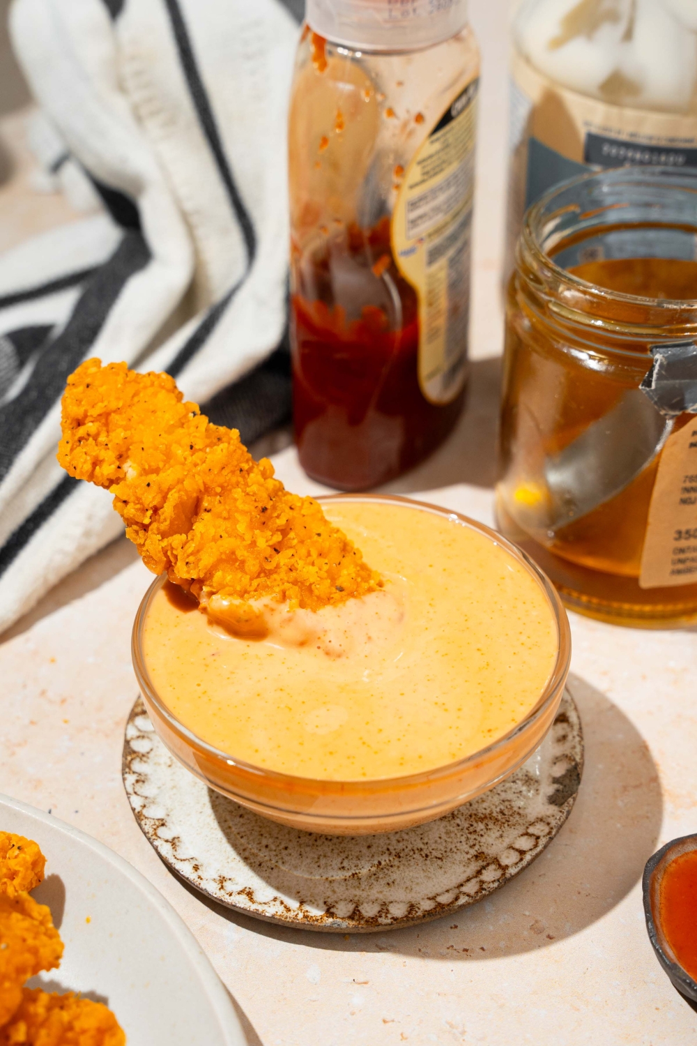A bowl of bang bang sauce on a saucer with a chicken tender dipped in the sauce. The saucer is on a tan counter with a jar of honey, and black striped napkin.