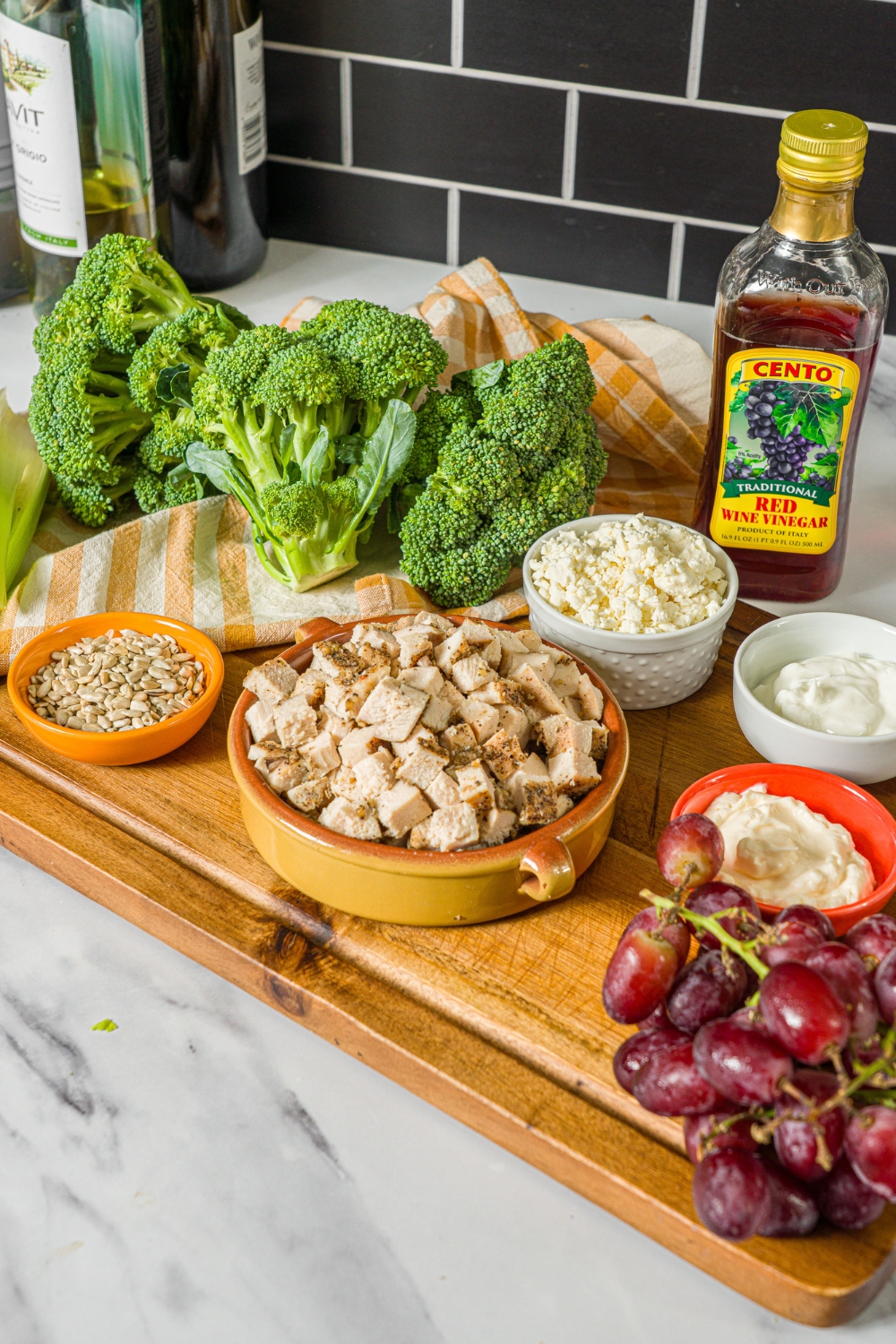 A wooden board with several bowls of ingredients to make broccoli chicken salad including broccoli, cubed chicken, grapes, celery, sunflower seeds, feta cheese, greek yogurt, and mayo. The board is on a marble counter.