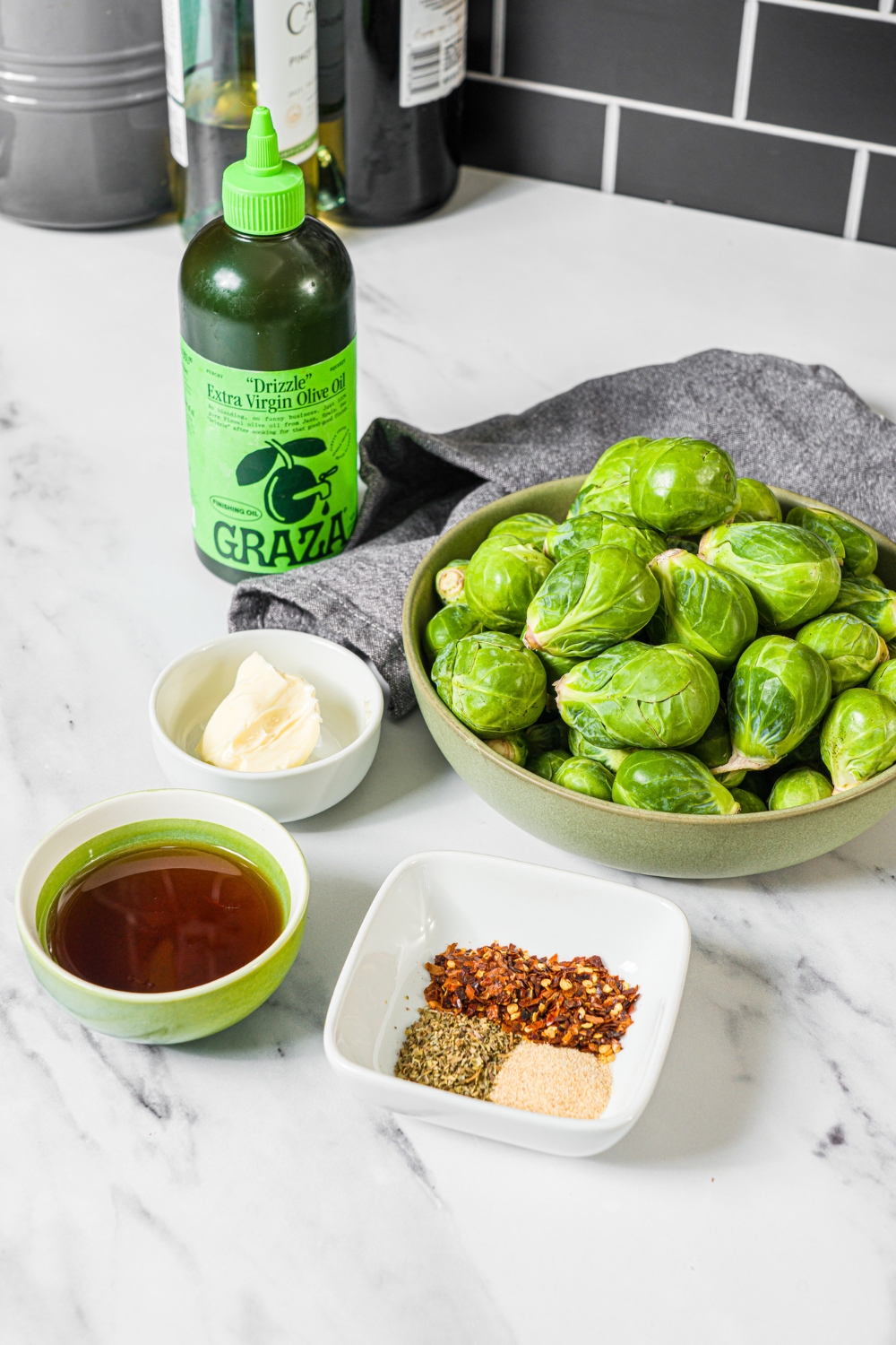 A marble counter with ingredients to make sweet chili brussels sprouts including brussels sprouts, oil, butter, maple syrup, red pepper flakes, and seasonings.
