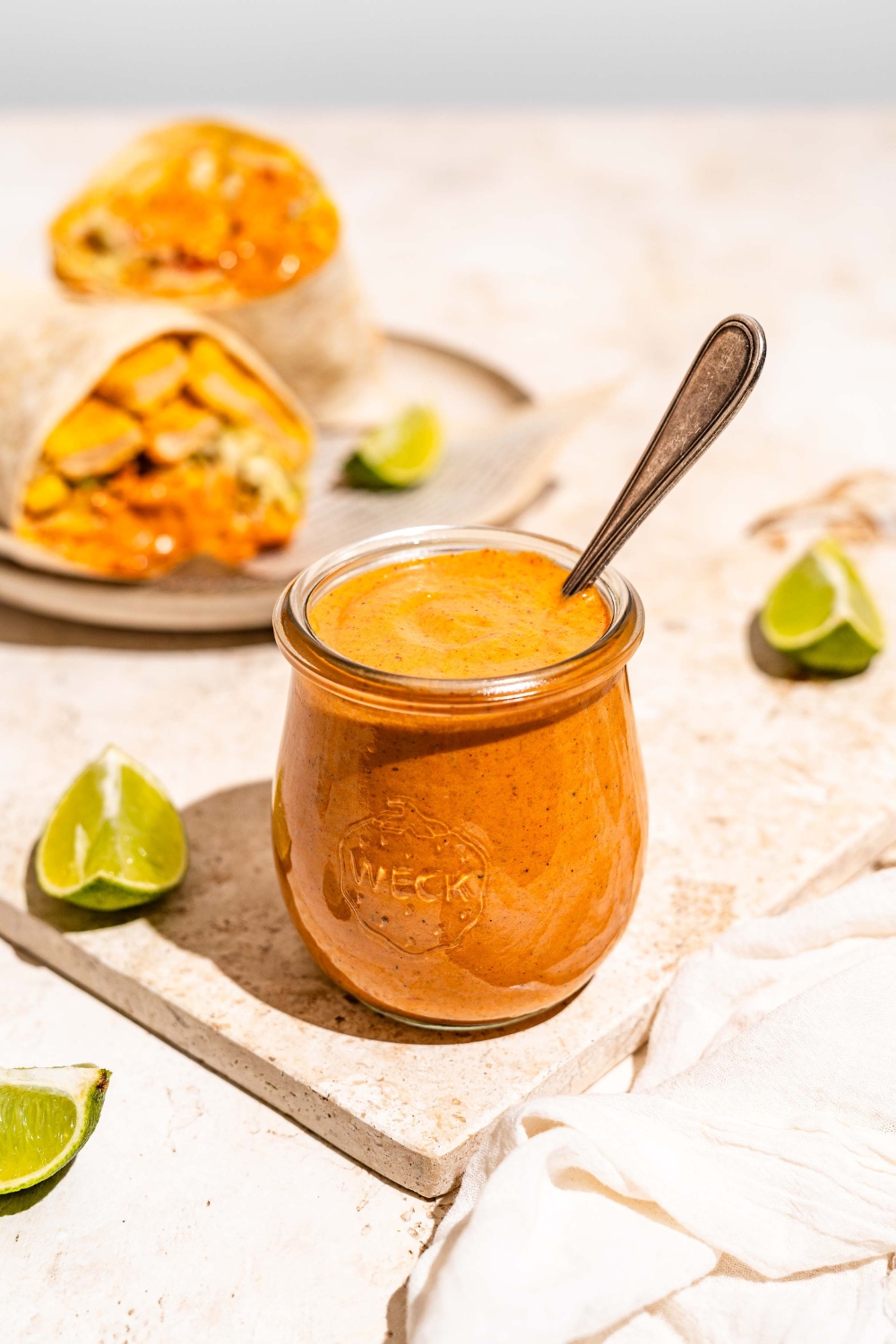 A jar of burrito sauce on a marble slab with lime wedges. There is a spoon in the jar. The slab is on a tan counter with a burrito on a plate.