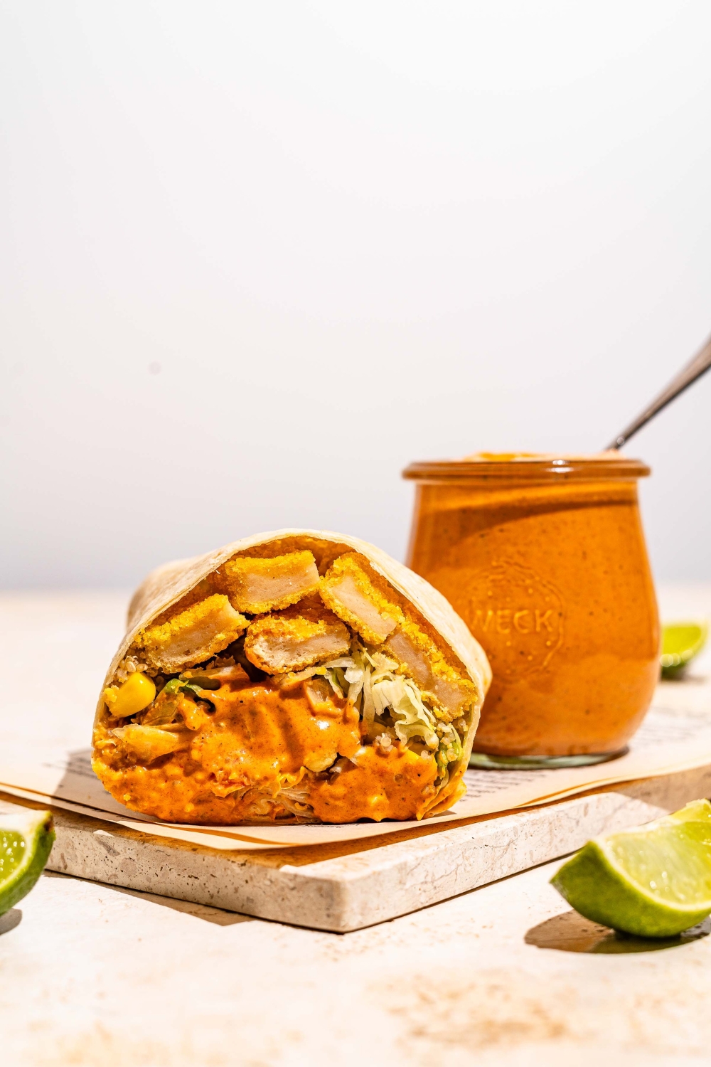 A burrito sliced in half and dipped in burrito sauce on a marble slab with a jar of burrito sauce. There is a spoon in the jar. The jar is on a tan counter with lime wedges.