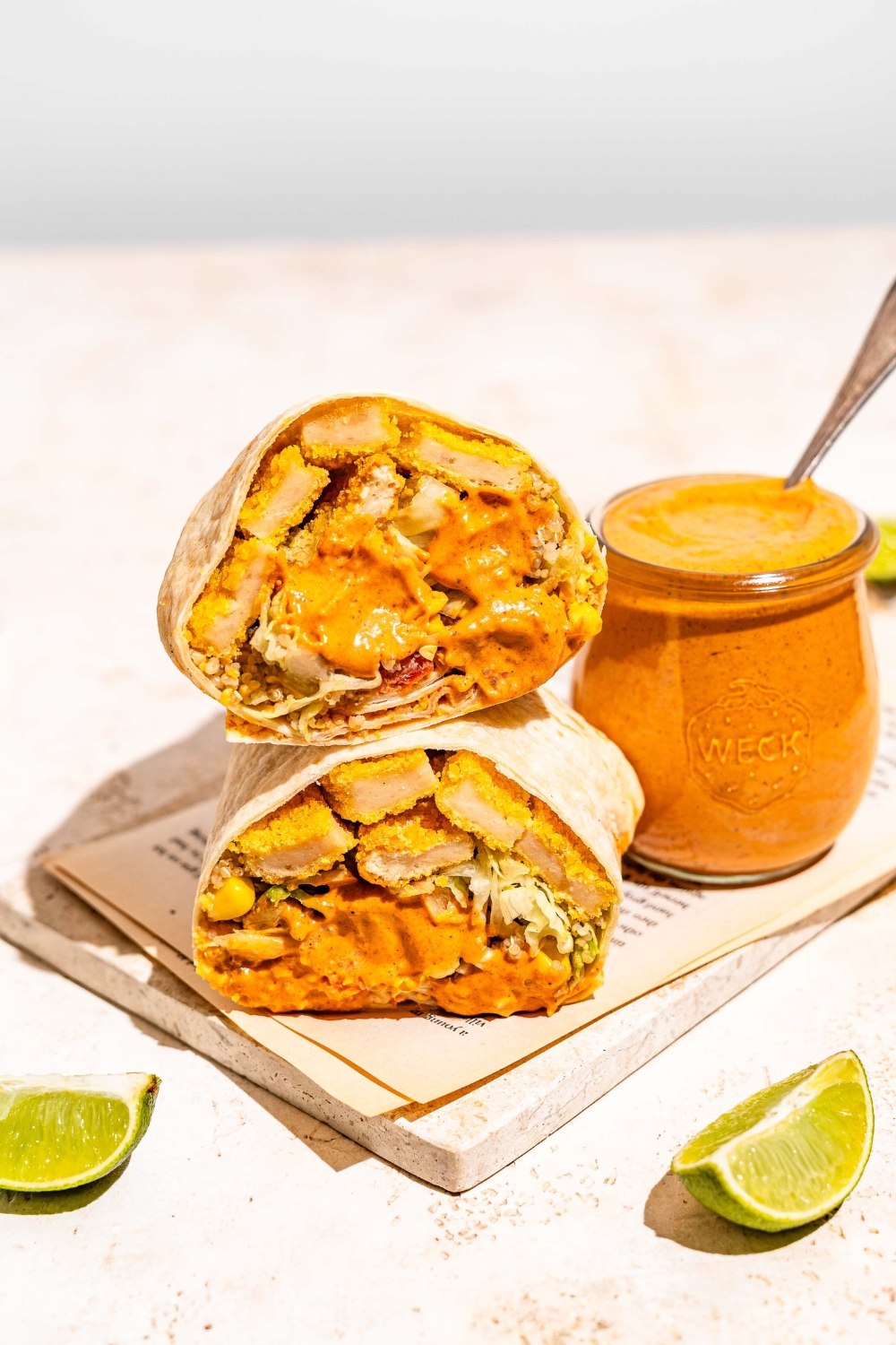 A burrito sliced in half and dipped in burrito sauce on a marble slab with a jar of burrito sauce. There is a spoon in the jar. The jar is on a tan counter with lime wedges.