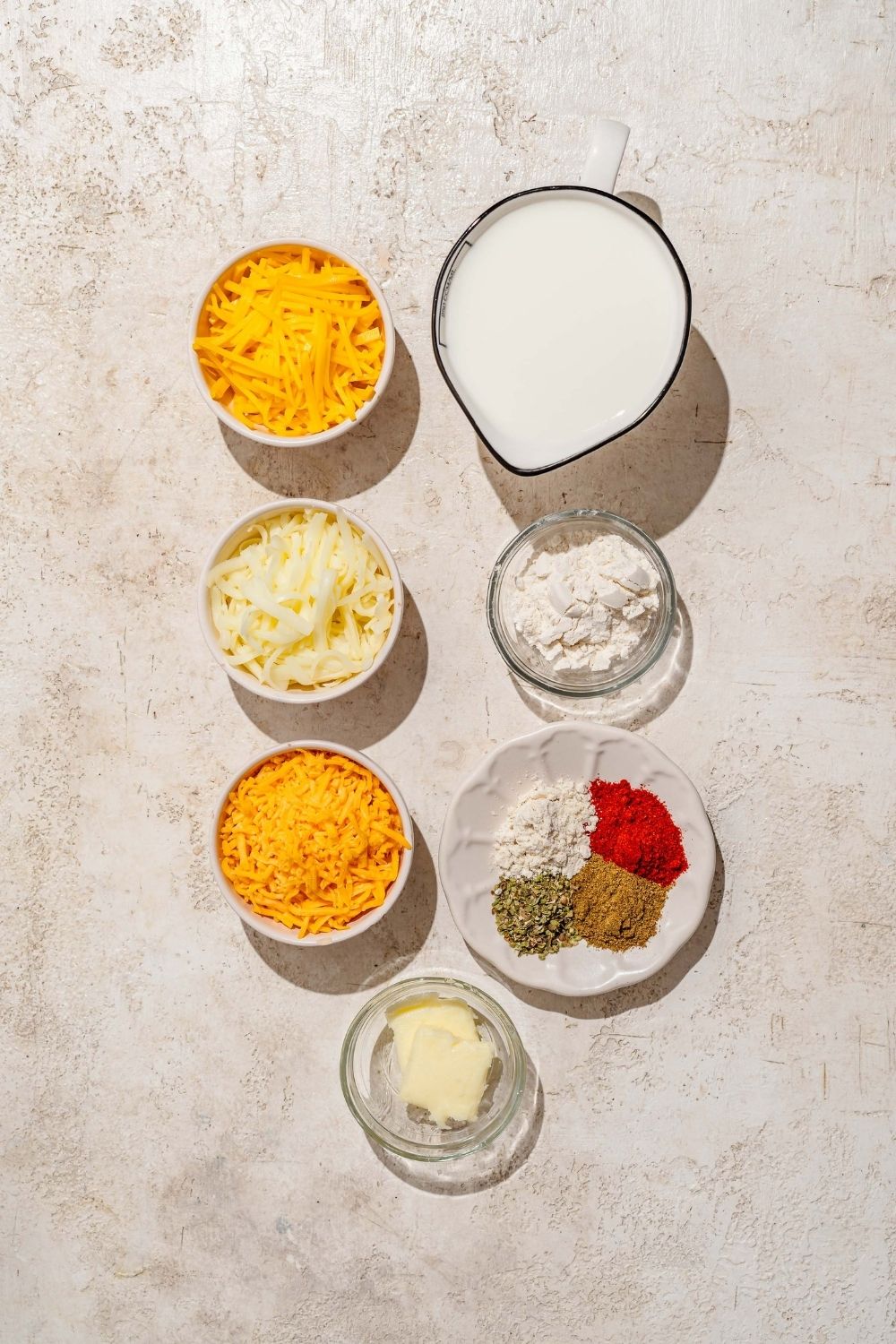 A tan counter with several bowls containing ingredients to make cheddar cheese sauce including shredded cheddar cheese, butter, milk, Monterey jack cheese, and seasonings.