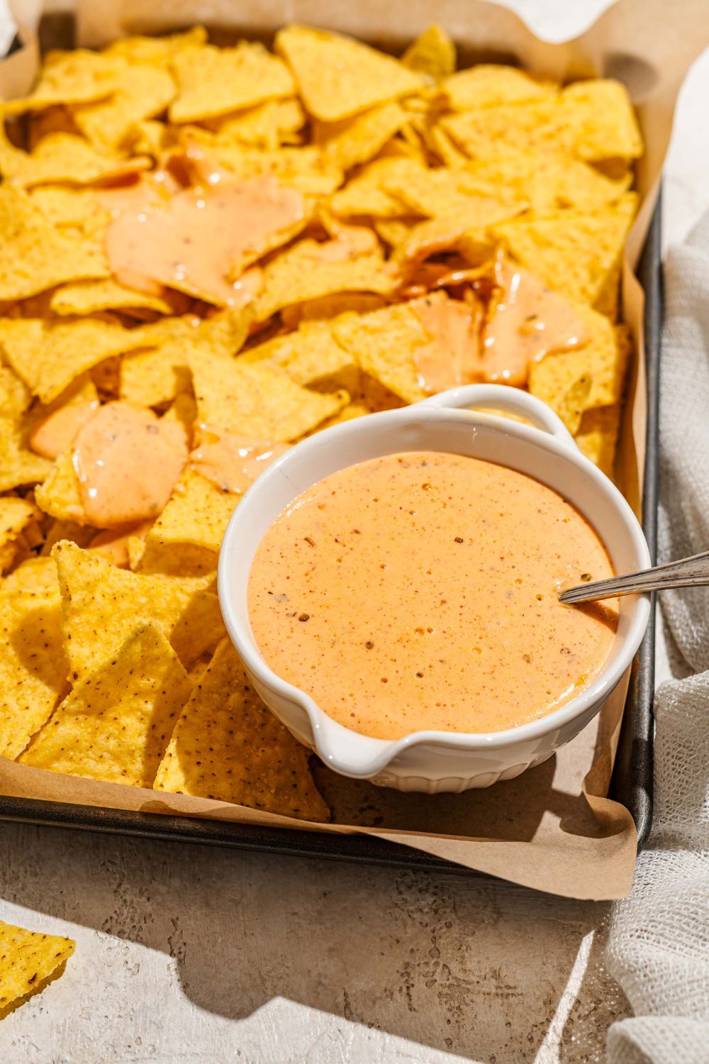 A baking sheet lined with parchment paper with nacho chips and a bowl of cheddar cheese sauce. A spoon is in the bowl.