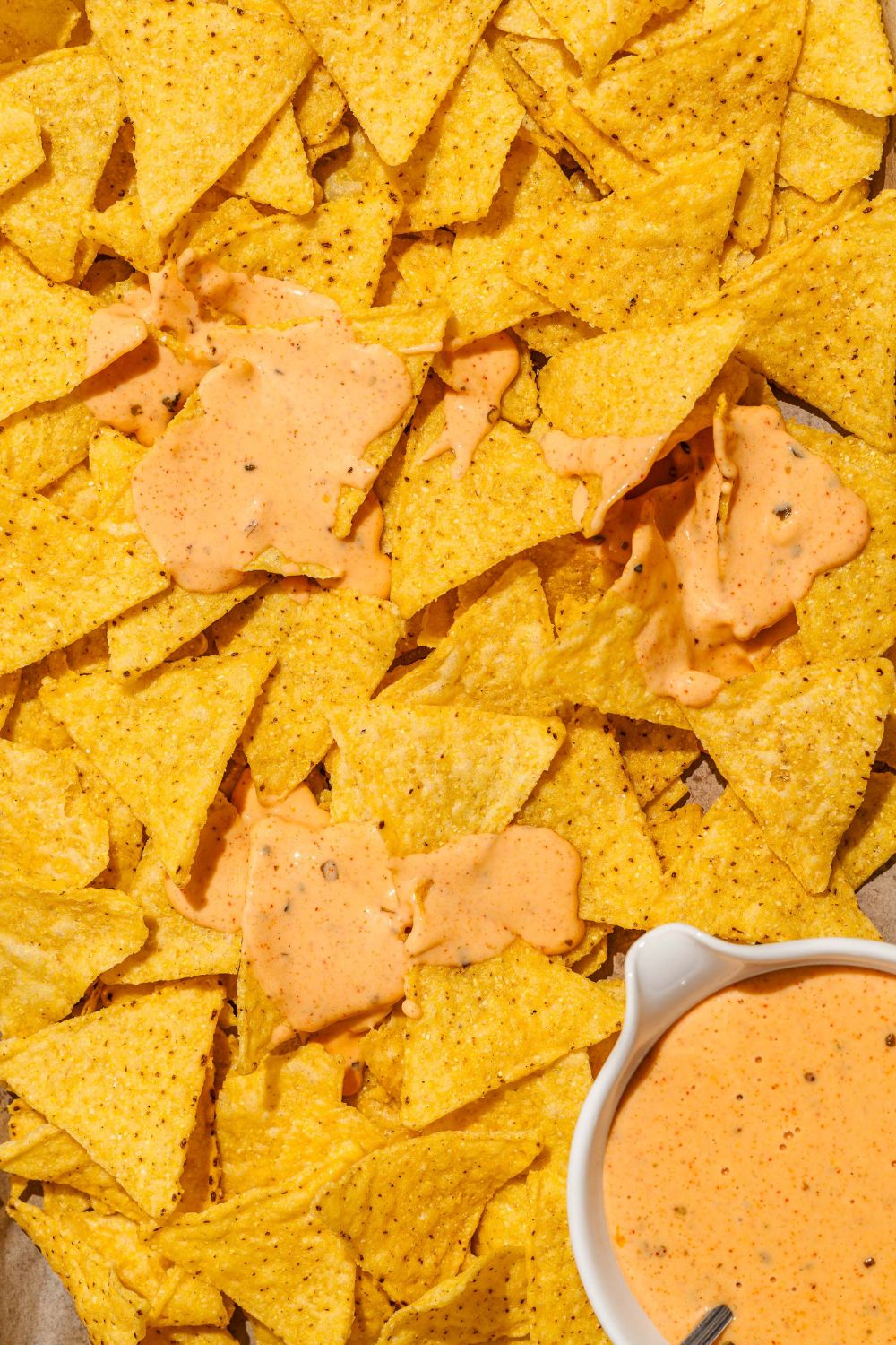 A baking sheet with nacho chips and a bowl of cheddar cheese sauce. The chips are drizzled with cheese sauce.
