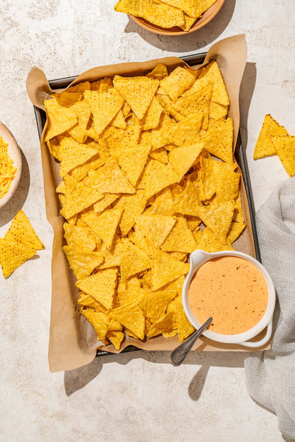 A baking sheet lined with parchment paper with nacho chips and a bowl of cheddar cheese sauce. A spoon is in the bowl. The sheet is on a tan counter with a bowl of chips.