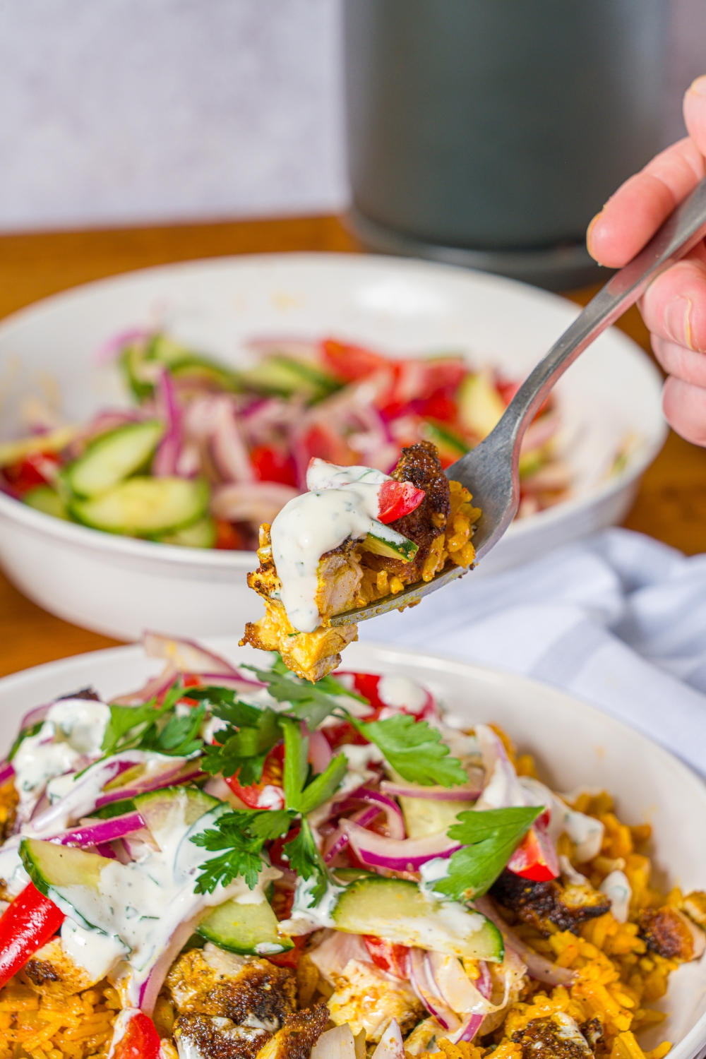 A white bowl with chicken shawarma bowl served with seasoned rice, chicken shawarma, onion salad, and garlic sauce. A fork is taking a bite from the bowl. The bowl is on a wooden counter with a bowl of onion salad.