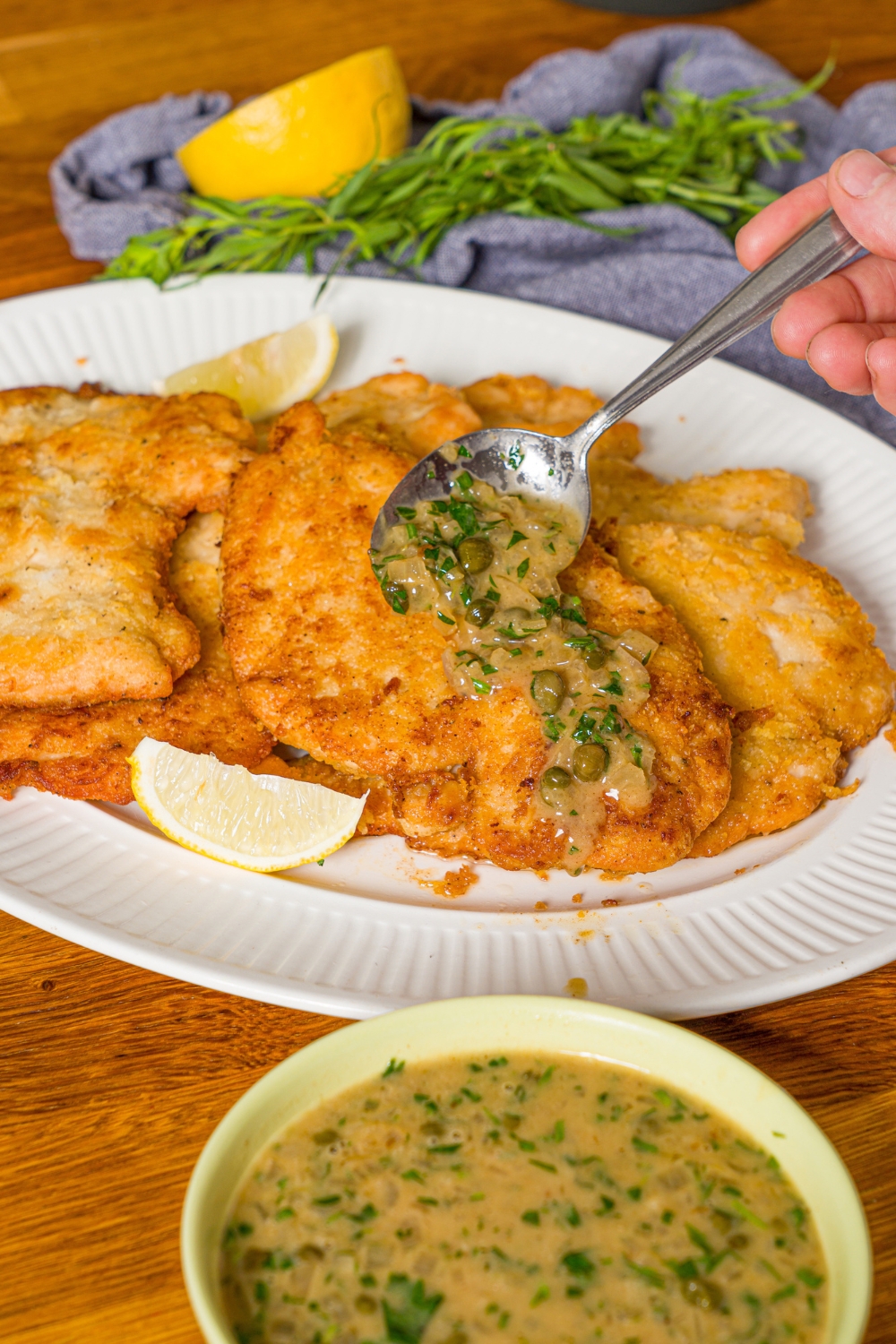 A white plate with chicken piccata sauce being poured over chicken piccata with a spoon. The plate is on a wooden counter with a bowl of piccata sauce.