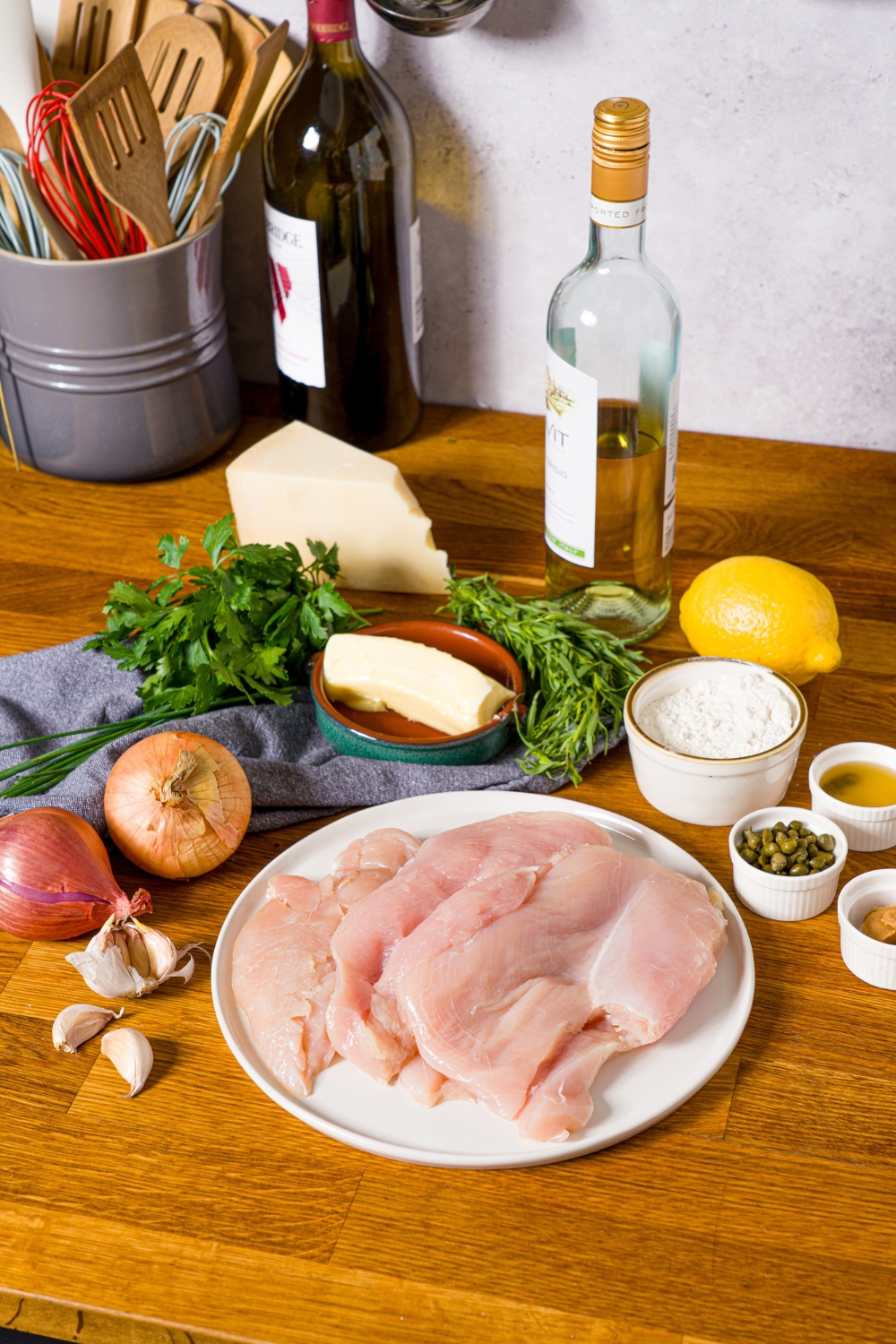 A wooden counter with ingredients to make chicken piccata sauce including chicken, onion, shallots, garlic, oil, white wine, butter, capers, lemon, and more.