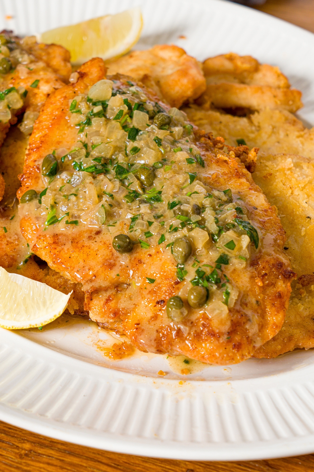 A white plate with chicken topped with chicken piccata sauce. The plate is served with lemon.
