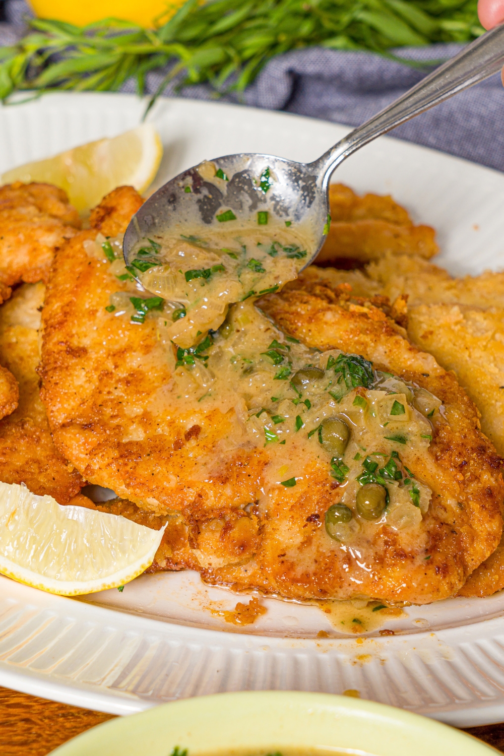 A white plate with chicken piccata sauce being poured over chicken piccata with a spoon. The plate is on a wooden counter.