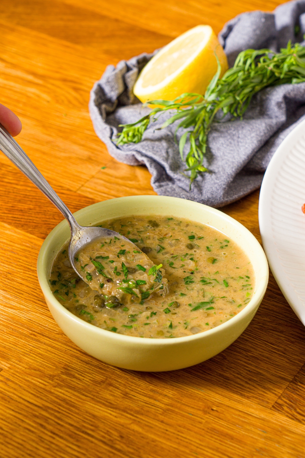 A bowl of chicken piccata sauce with a spoon dipping in the bowl. The bowl is on a wooden counter with a lemon and blue cloth napkin.