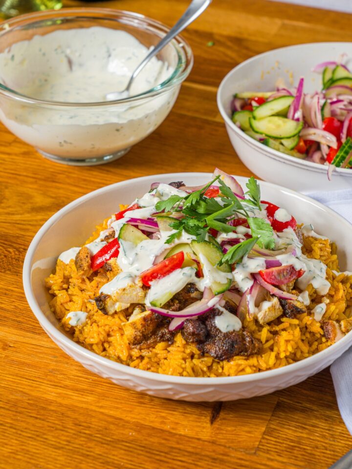 A white bowl with chicken shawarma bowl served with seasoned rice, chicken shawarma, onion salad, and garlic sauce garnished with fresh parsley. The bowl is on a wooden counter with a bowl of onion salad and bowl of garlic sauce.
