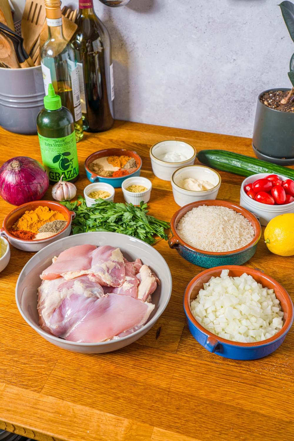 A wooden counter with several bowls containing ingredients to make chicken shawarma including chicken thighs, white onion, rice, olive oil, red onion, tomatoes, lemon, cucumber, garlic, greek yogurt, mayo, and seasonings.