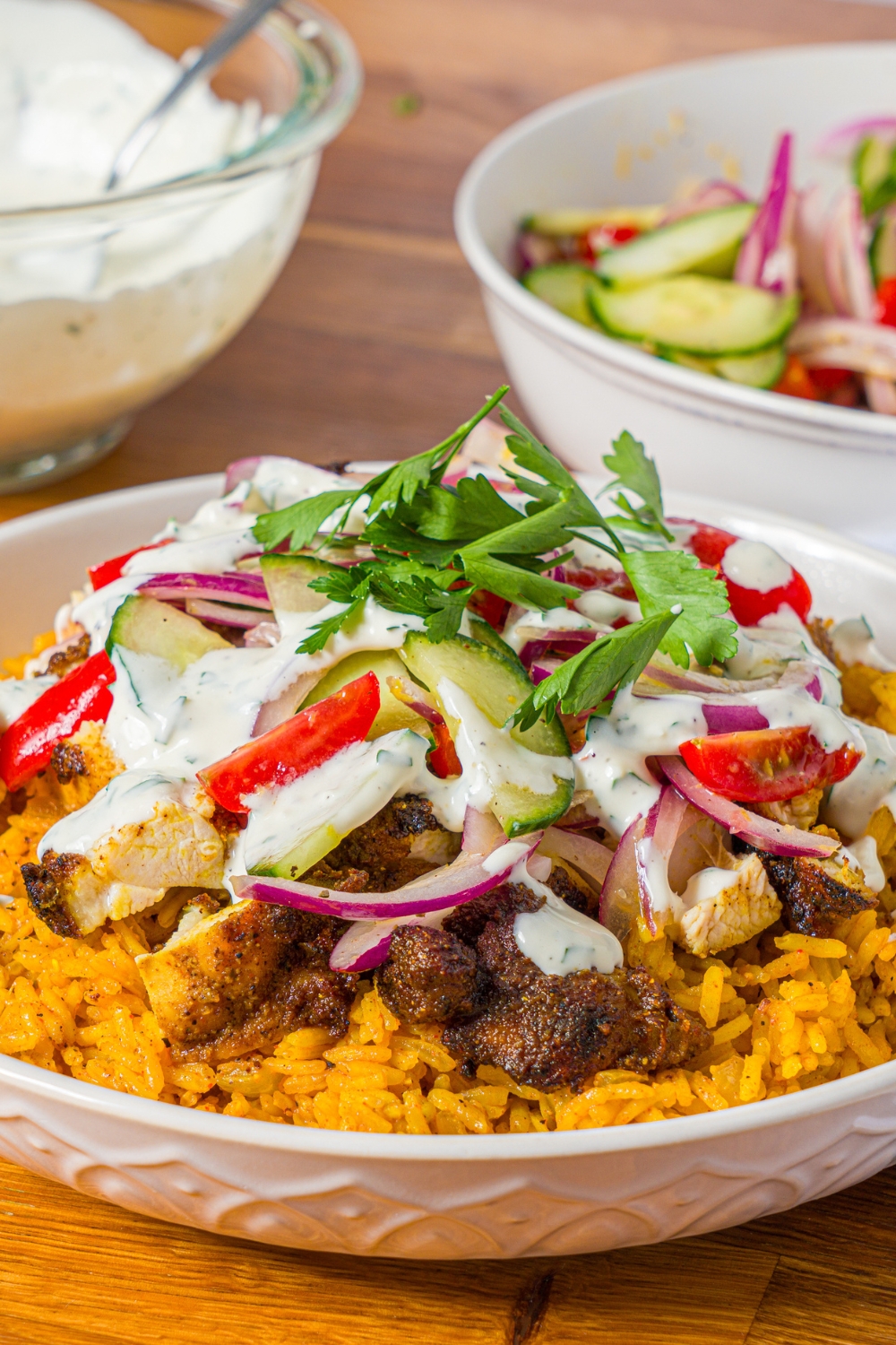 A white bowl with chicken shawarma bowl served with seasoned rice, chicken shawarma, onion salad, and garlic sauce garnished with fresh parsley. The bowl is on a wooden counter with a bowl of onion salad and bowl of garlic sauce.