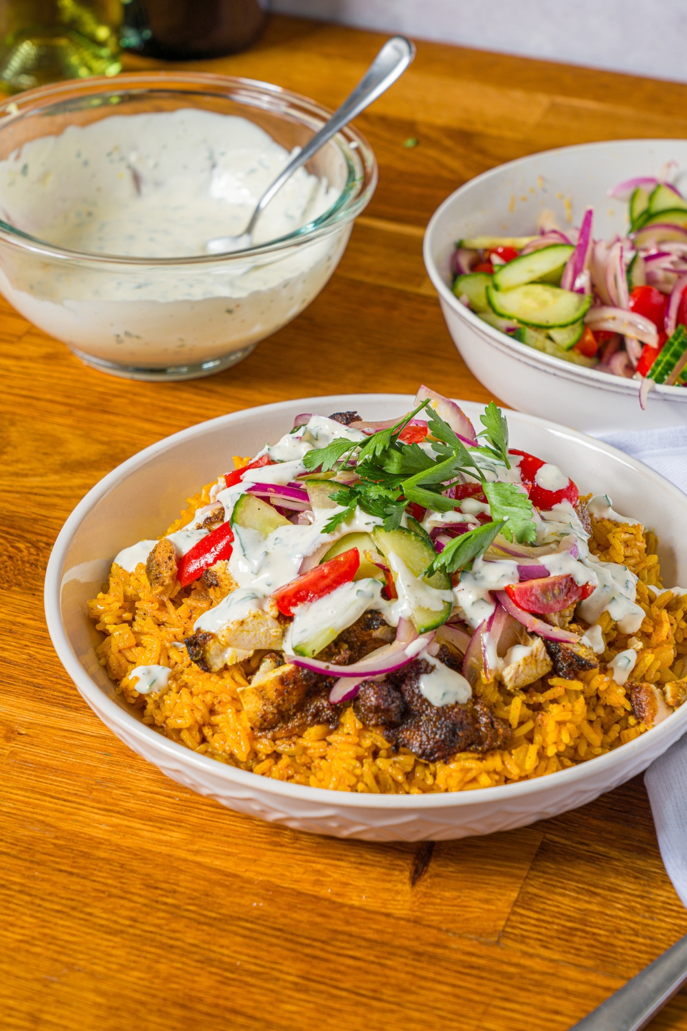 A white bowl with chicken shawarma bowl served with seasoned rice, chicken shawarma, onion salad, and garlic sauce garnished with fresh parsley. The bowl is on a wooden counter with a bowl of onion salad and bowl of garlic sauce.