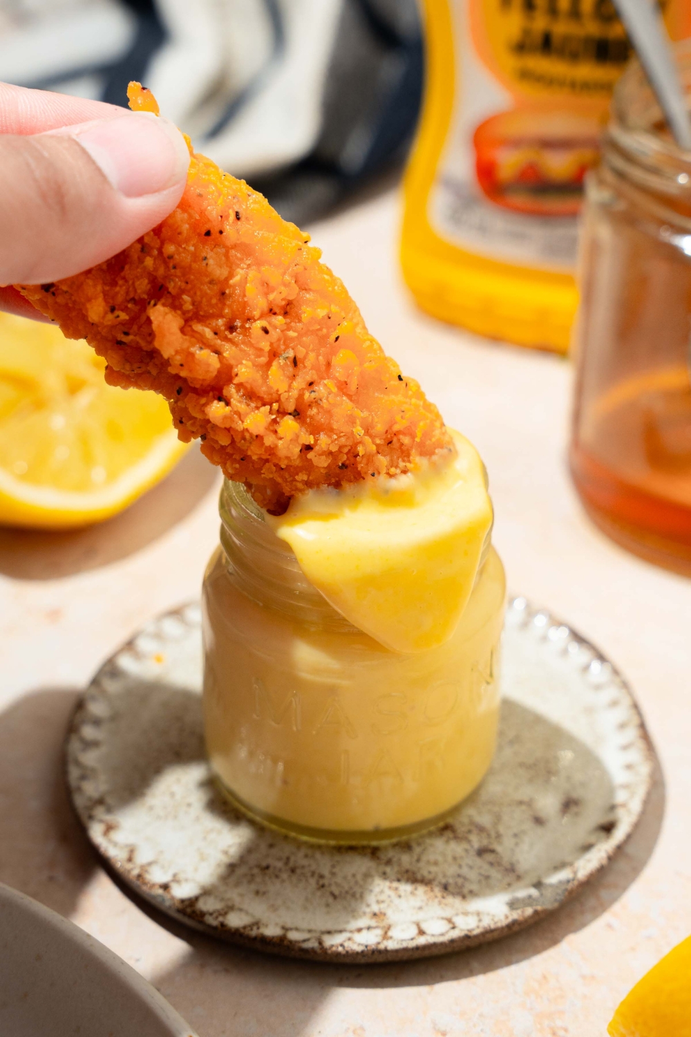 A jar of honey mustard sauce with a chicken tender dipping into the jar. The jar is on a saucer on a tan counter with a jar of honey and lemon.