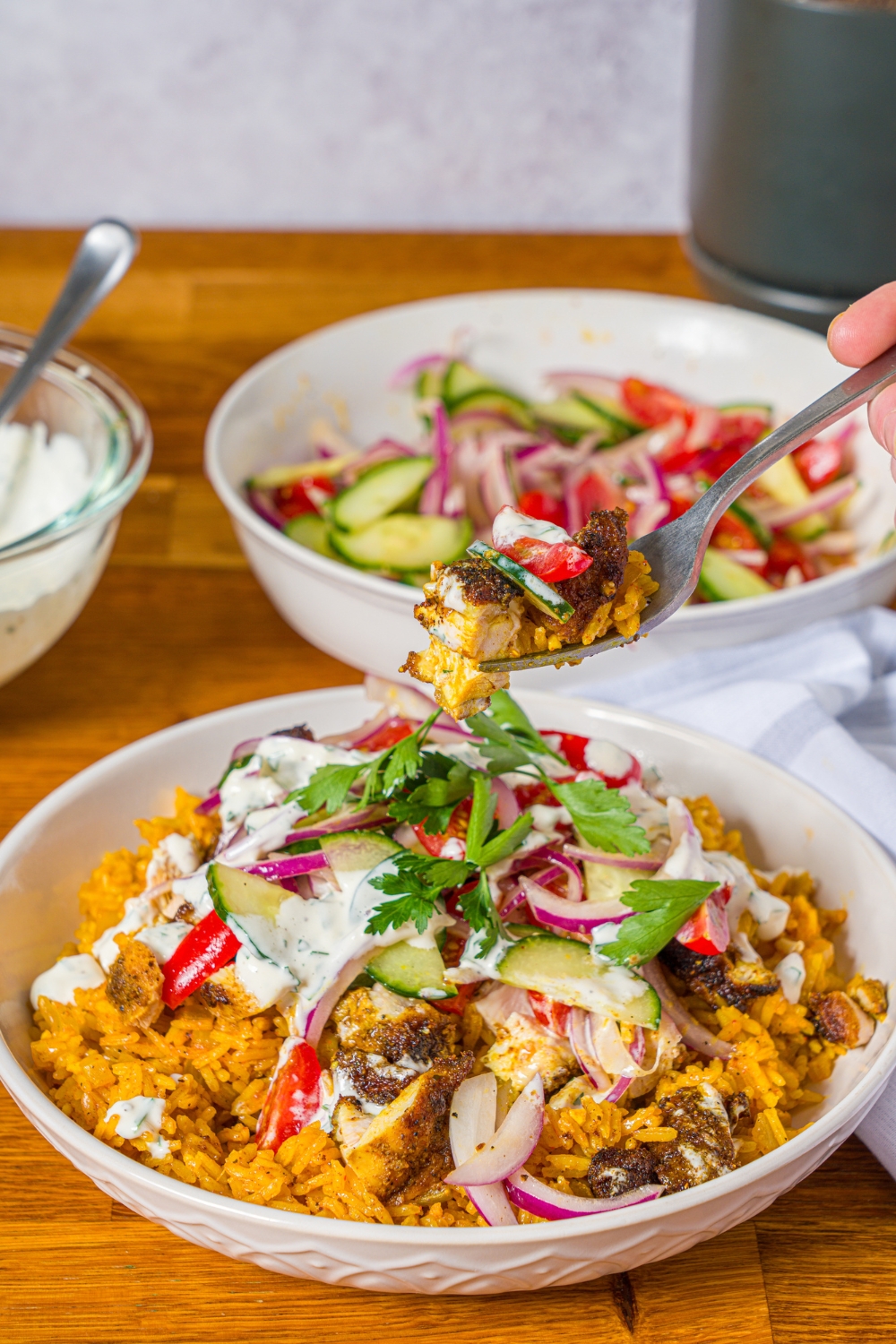 A white bowl with chicken shawarma bowl served with seasoned rice, chicken shawarma, onion salad, and garlic sauce garnished with fresh parsley. A fork is taking a bite from the bowl. The bowl is on a wooden counter with a bowl of onion salad and bowl of garlic sauce.