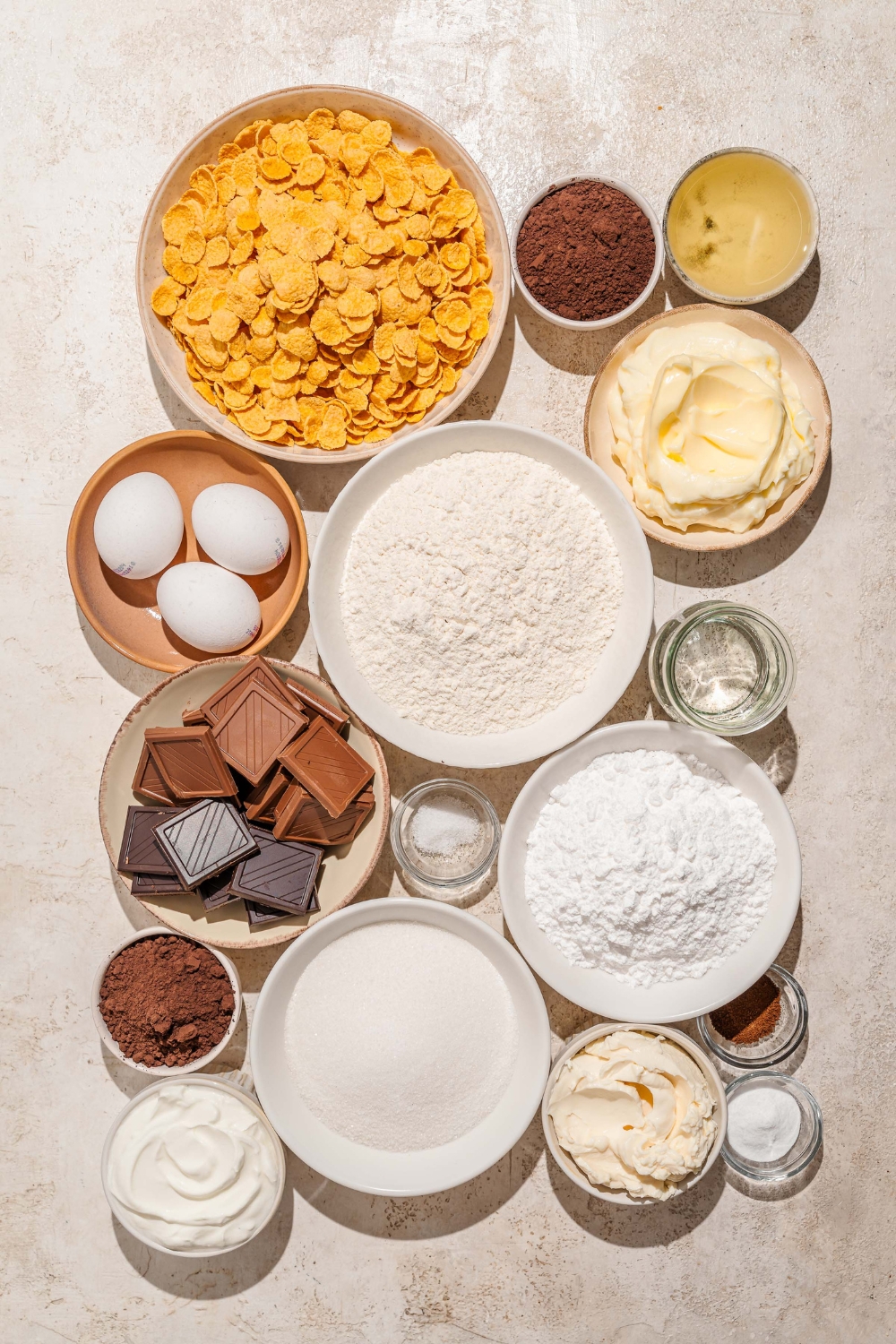 A tan counter with several bowls of ingredients to make chocolate crunch cake including cornflakes, flour, eggs, butter, chocolate, cocoa powder, cream cheese, and more.