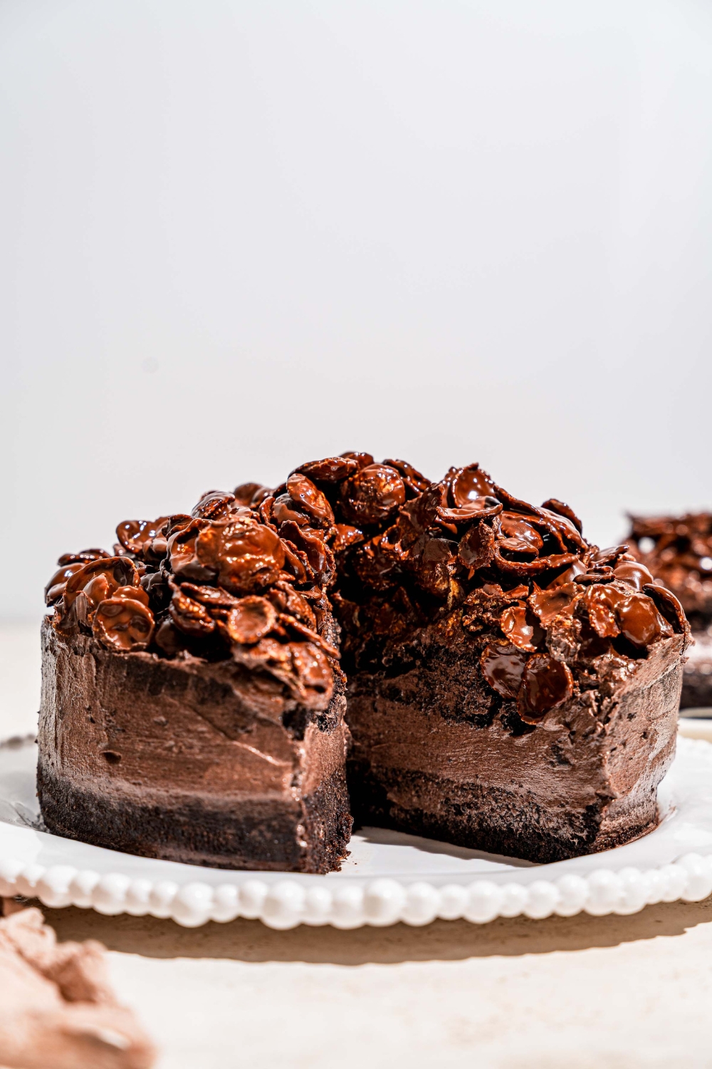 A white platter with a whole chocolate crunch cake topped with chocolate cornflake crunch. A slice is removed from the cake. The platter is on a tan counter.