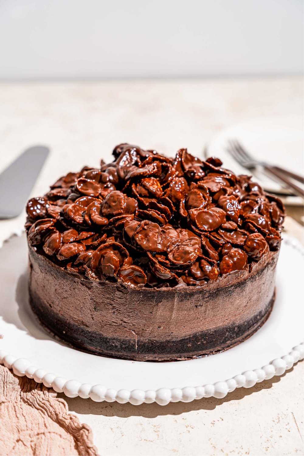 A white platter with a whole chocolate crunch cake topped with chocolate cornflake crunch. The platter is on a tan counter.