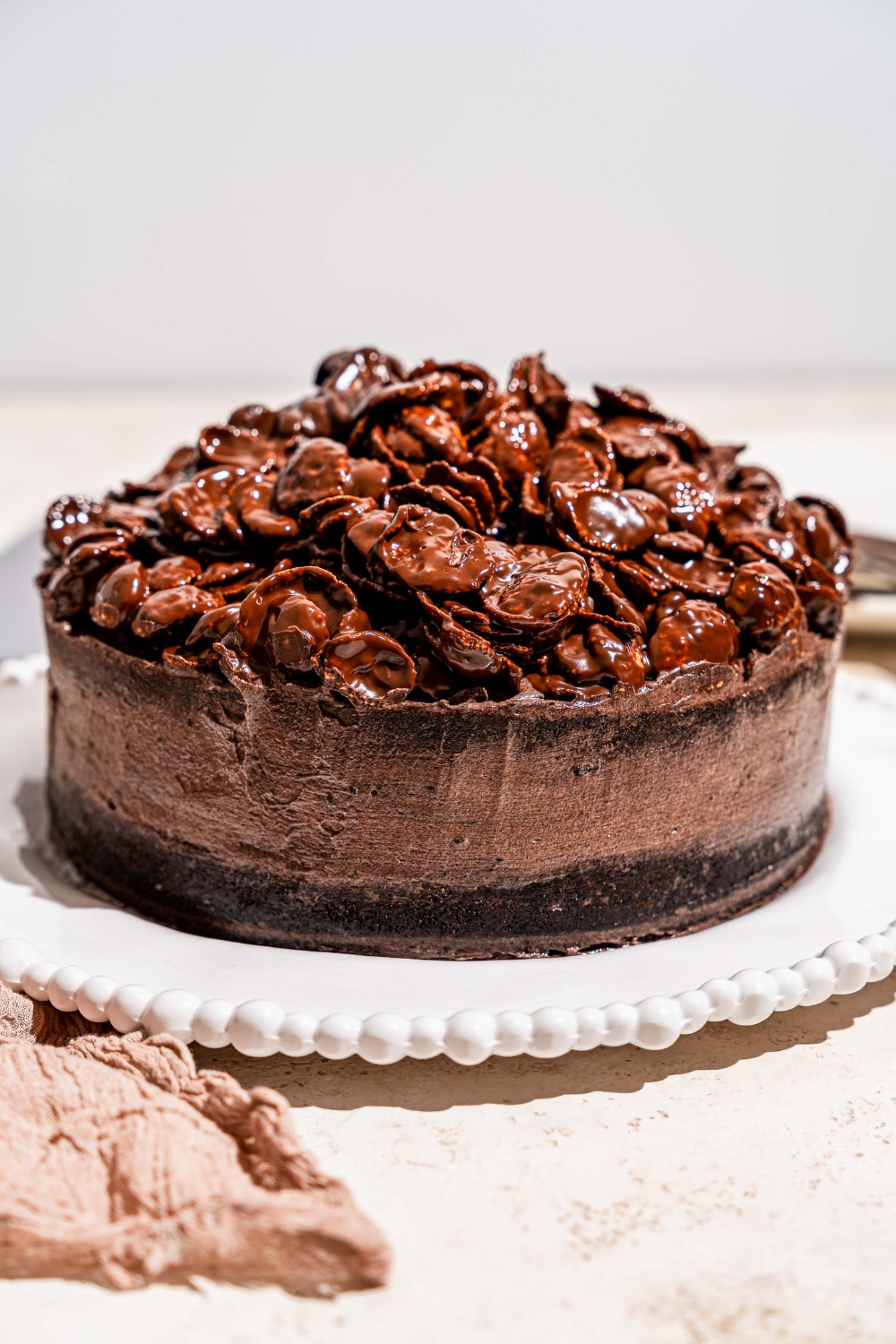 A white platter with a whole chocolate crunch cake topped with chocolate cornflake crunch. The platter is on a tan counter with a plate with a slice of cake.