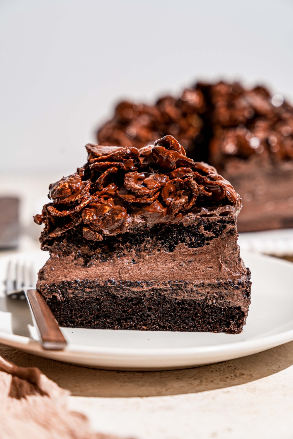 A white plate with a slice of chocolate crunch cake topped with chocolate cornflake crunch. The plate is on a tan counter with a platter of cake.
