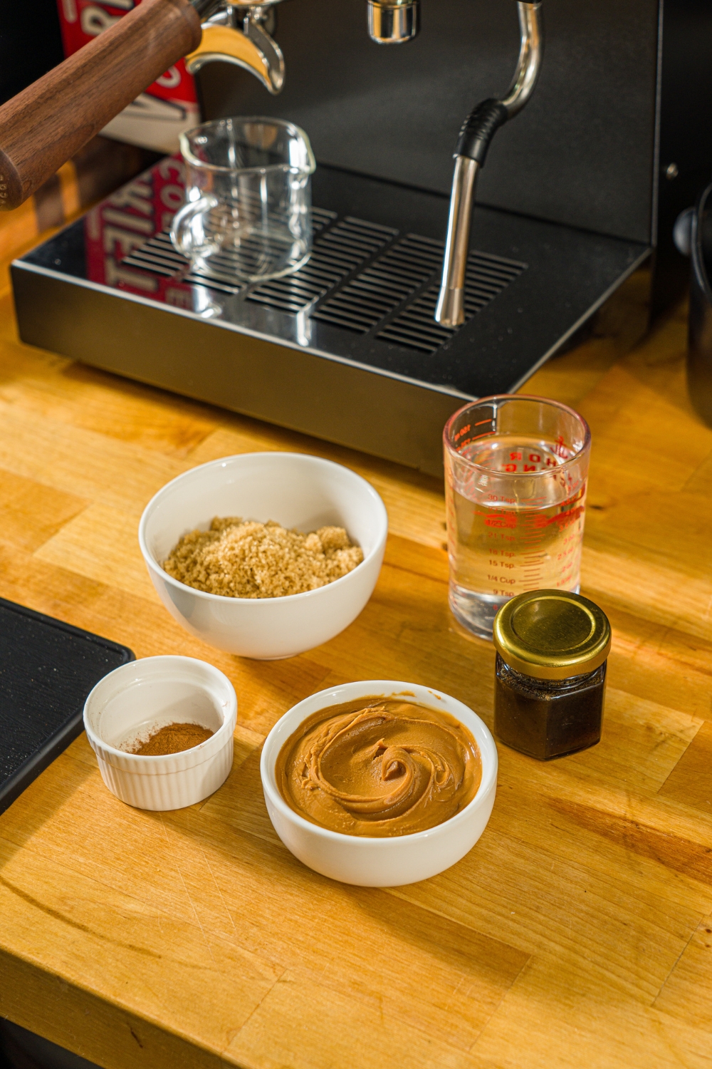 A wooden counter with several bowls of ingredients to make cookie butter latte including brown sugar, cookie butter, vanilla paste, and water.