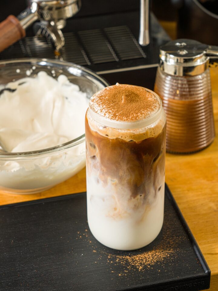 A tall glass with iced cookie butter latte topped with cold foam and cinnamon. The glass is on a wooden counter with a bowl of cold foam and an espresso machine.