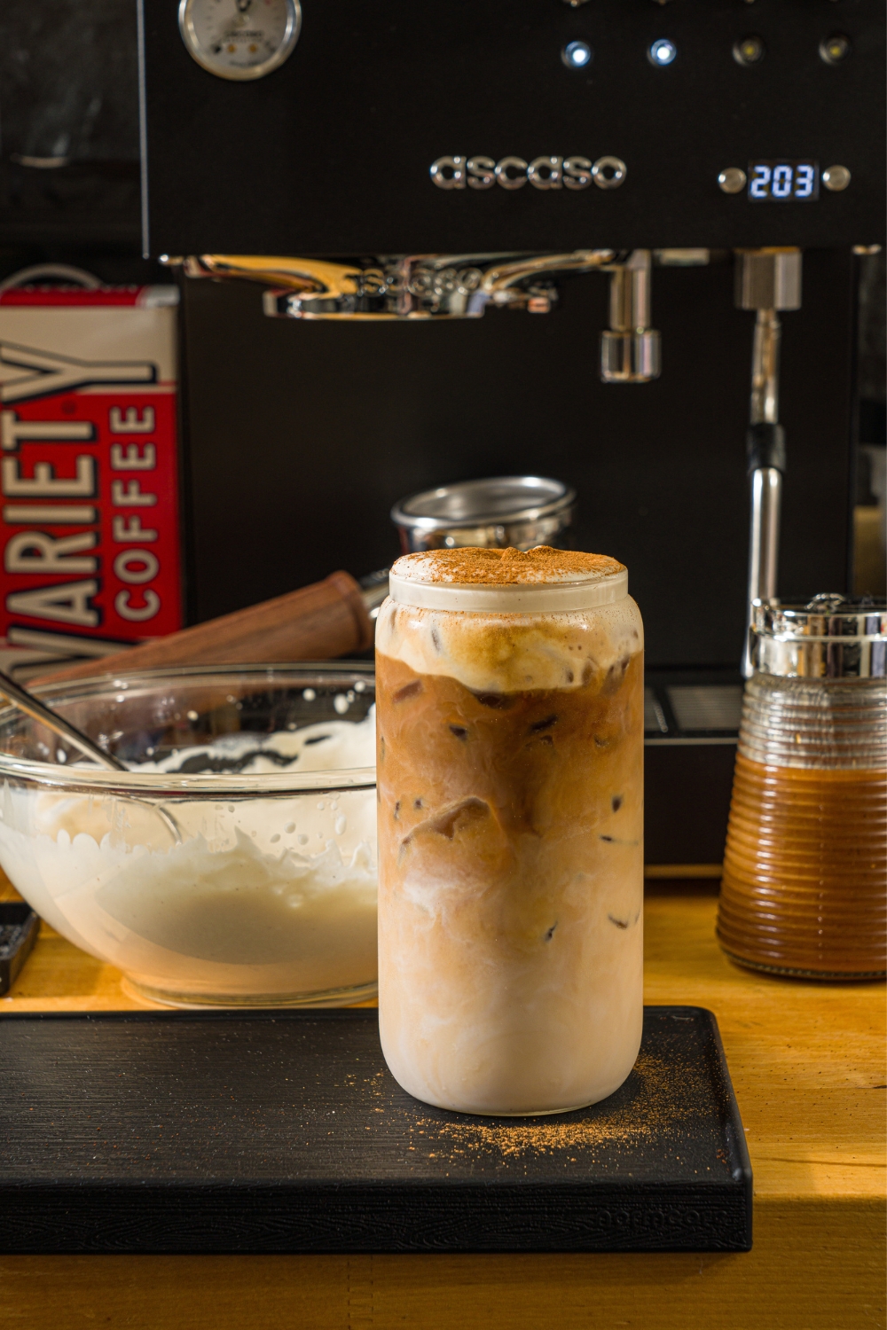 A tall glass with iced cookie butter latte topped with cold foam and cinnamon. The glass is on a wooden counter with a bowl of cold foam and an espresso machine.