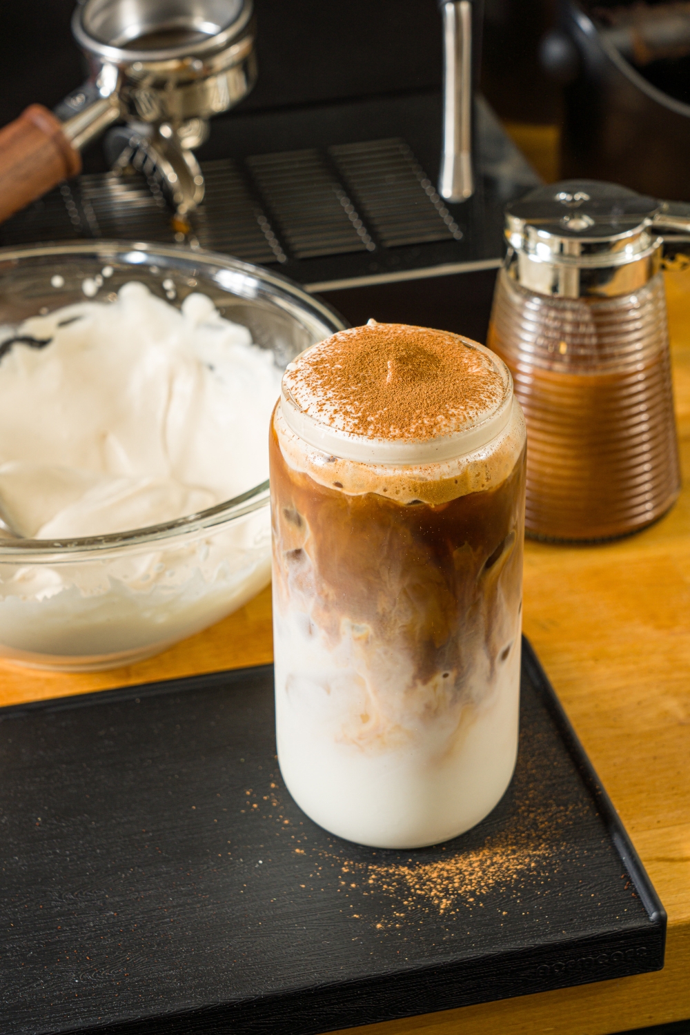 A tall glass with iced cookie butter latte topped with cold foam and cinnamon. The glass is on a wooden counter with a bowl of cold foam and an espresso machine.