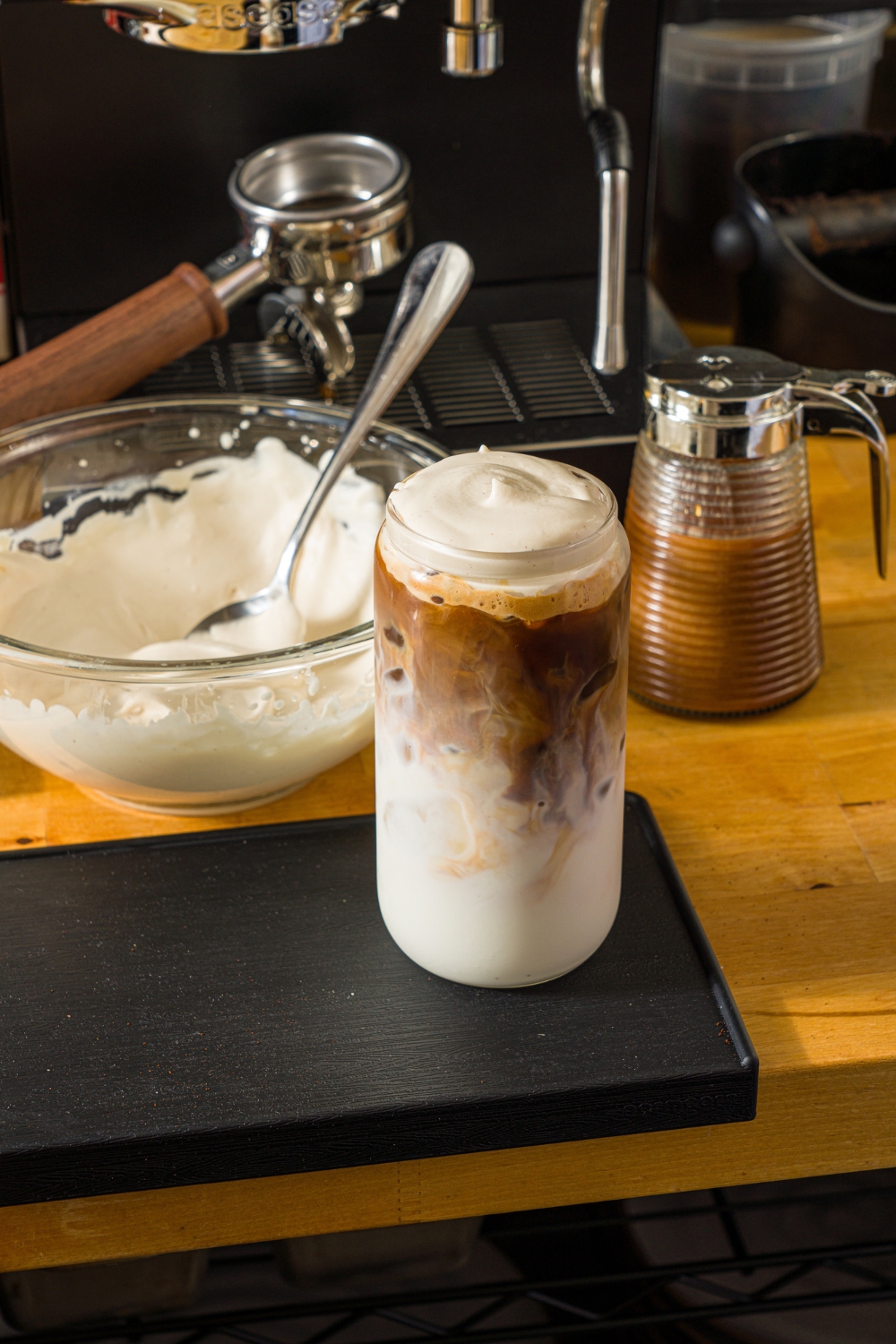 A tall glass with iced cookie butter latte topped with cold foam and cinnamon. The glass is on a wooden counter with a bowl of cold foam and an espresso machine.
