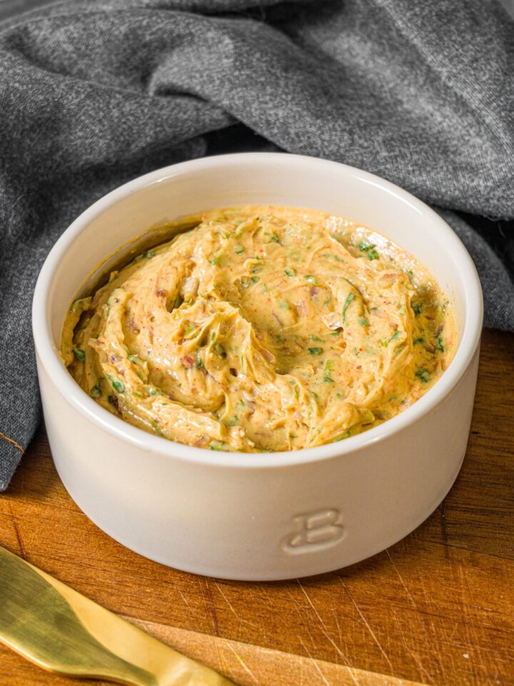 A small bowl of cowboy butter on a wooden board with a butter knife and grey cloth napkin.