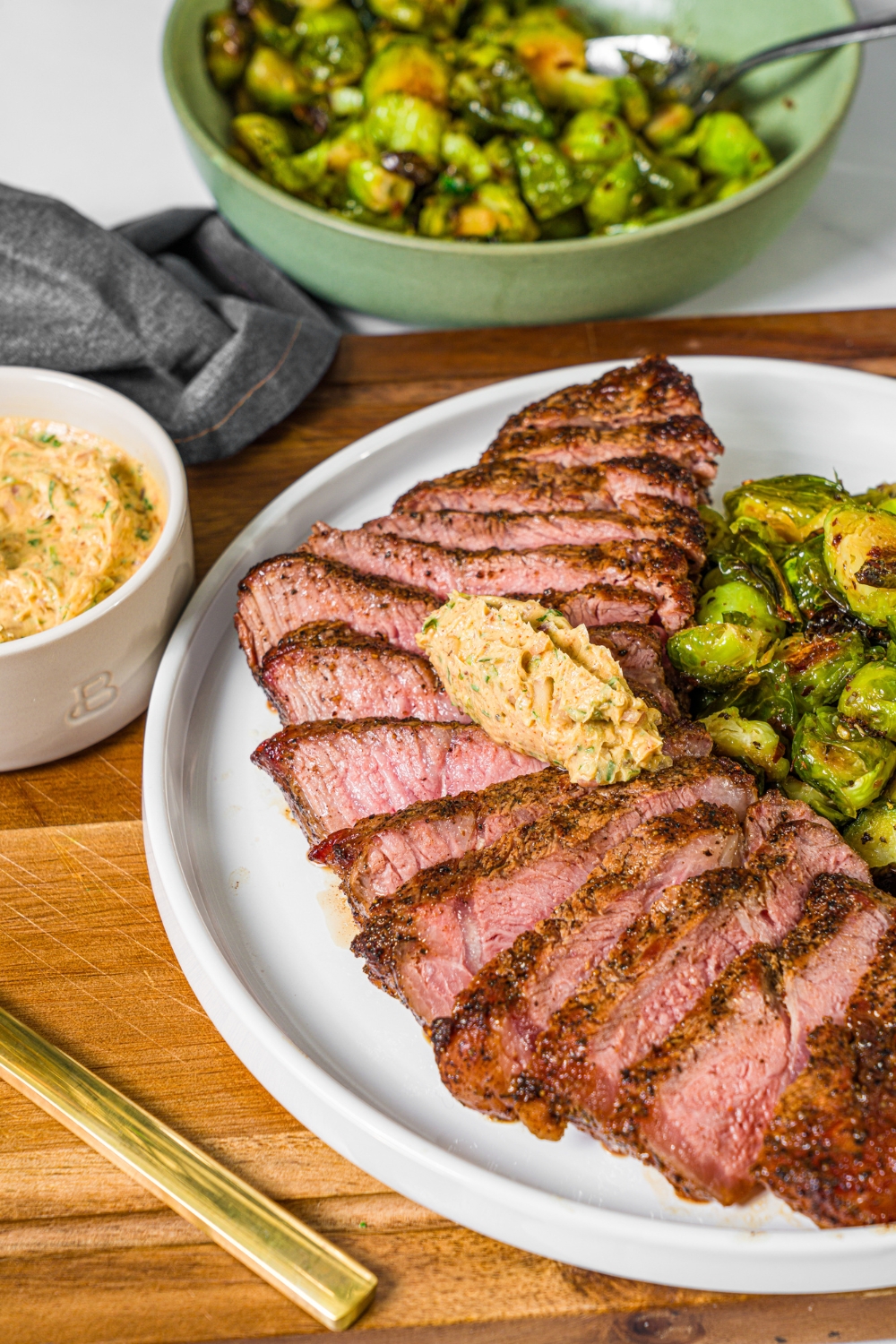A white plate with sliced grilled steak topped with cowboy butter and a side of roasted brussels sprouts. The plate is on a wooden board with a bowl of cowboy butter.