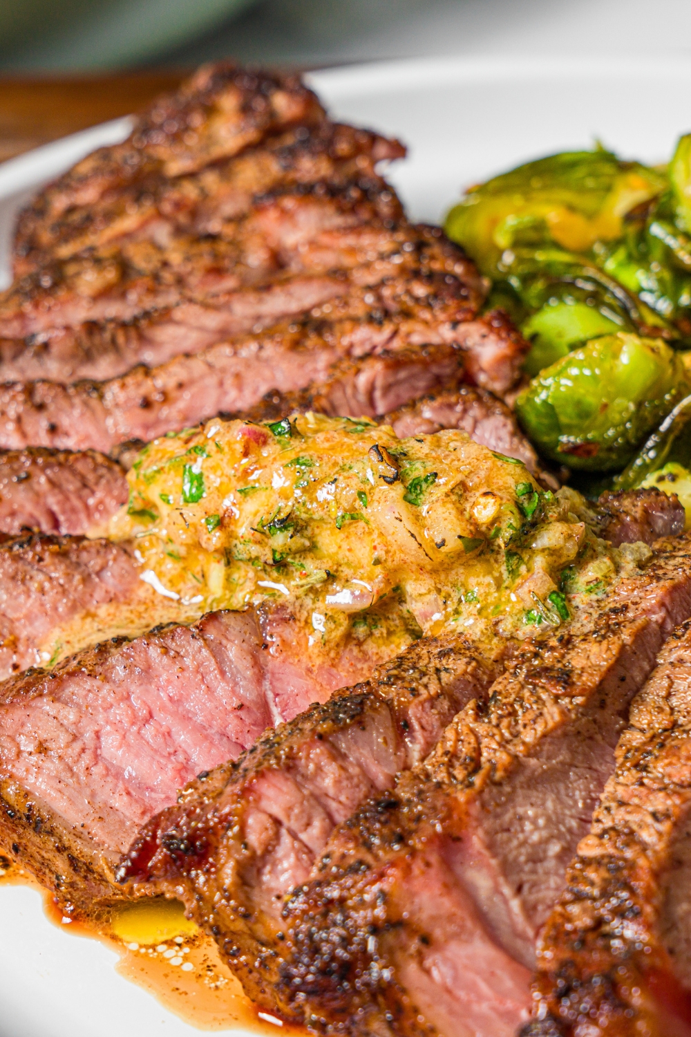 A white plate with sliced grilled steak topped with cowboy butter and served with roasted brussels sprouts.