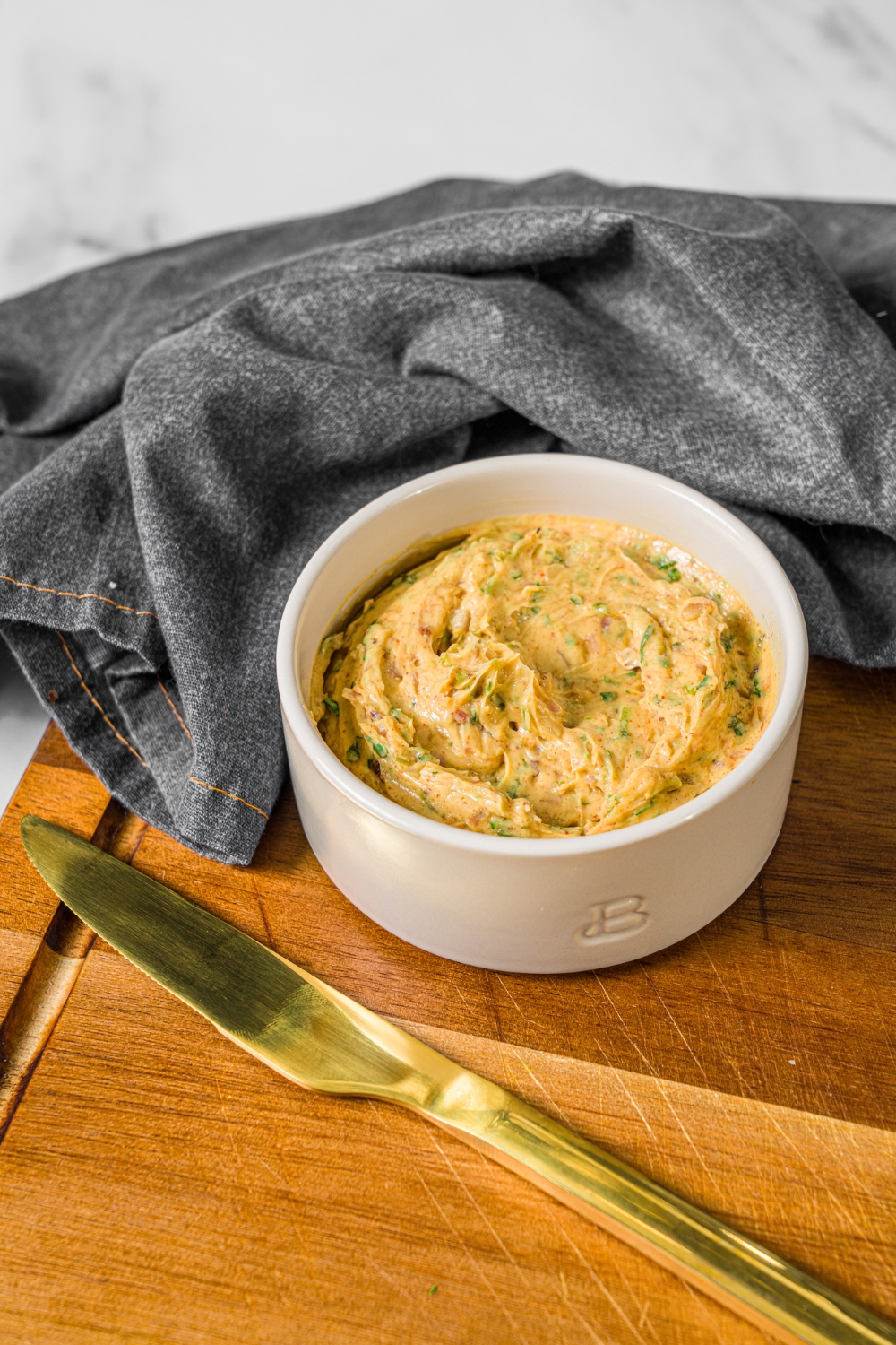 A small bowl of cowboy butter on a wooden board with a butter knife and grey cloth napkin.