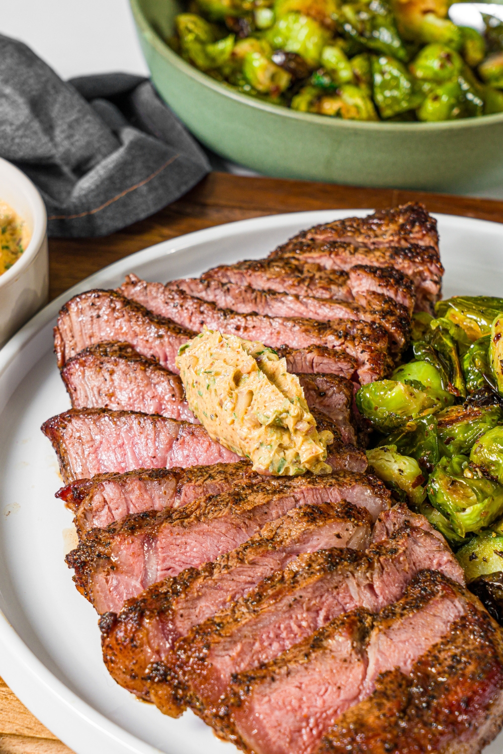 A white plate with sliced grilled steak topped with cowboy butter and a side of roasted brussels sprouts. The plate is on a wooden board with a bowl of brussels sprouts.