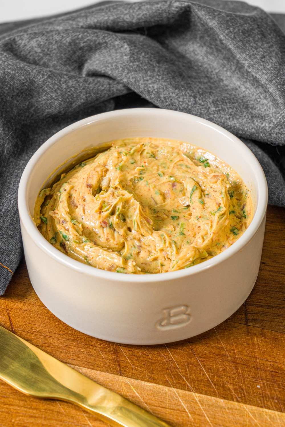 A small bowl of cowboy butter on a wooden board with a butter knife and grey cloth napkin.