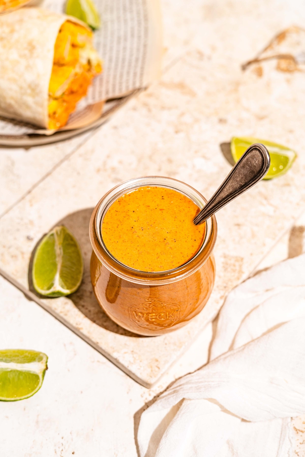 A jar of burrito sauce on a marble slab with lime wedges. There is a spoon in the jar. The slab is on a tan counter with a burrito on a plate.